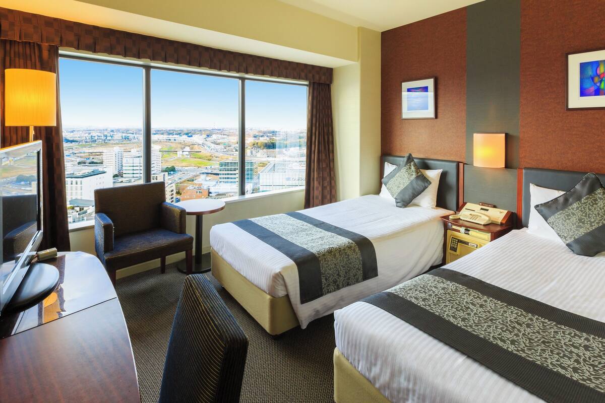 Ein Hotelzimmer mit zwei Betten, einem Schreibtisch, einem Stuhl, einem kleinen Tisch und einem großen Fenster mit Blick auf die Stadt.