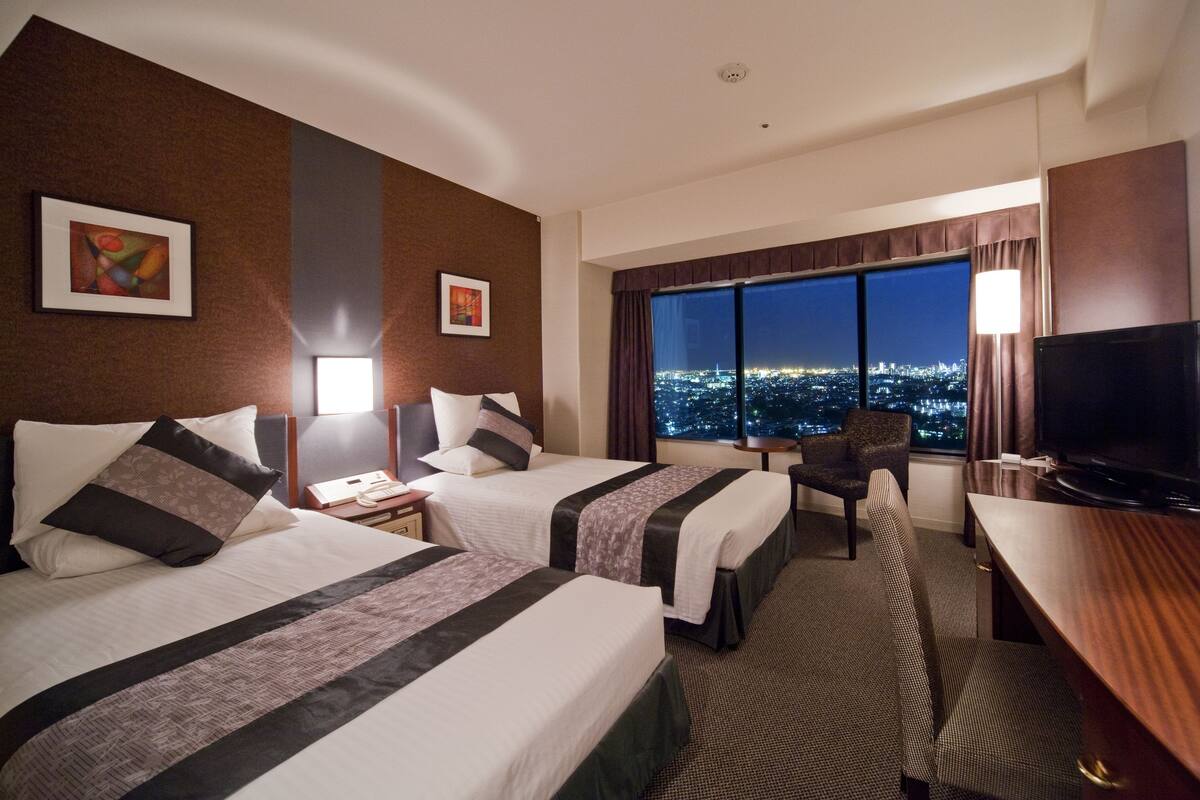 Ein Hotelzimmer mit zwei Betten, einem Schreibtisch, einem Sessel, einem Fernseher und einem großen Fenster mit Blick auf die Stadt.