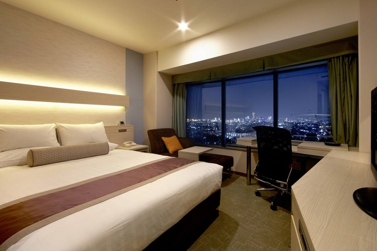 Ein Hotelzimmer mit einem großen Bett, einem Schreibtisch, einem Stuhl und Blick auf eine nächtliche Skyline.