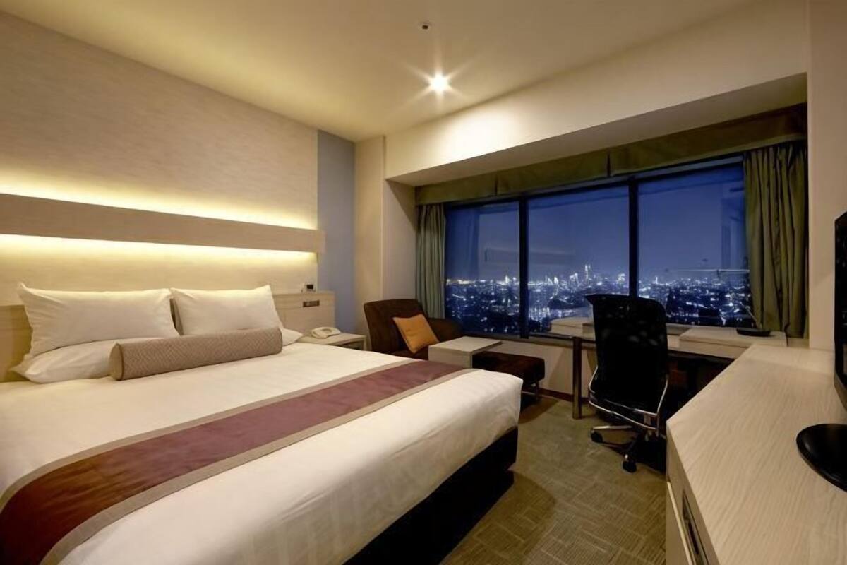 Ein Hotelzimmer mit einem großen Bett, einem Schreibtisch mit Stuhl und Blick auf eine Stadtlandschaft durch ein Fenster, sowie einem durch das Fenster sichtbaren Nachthimmel.