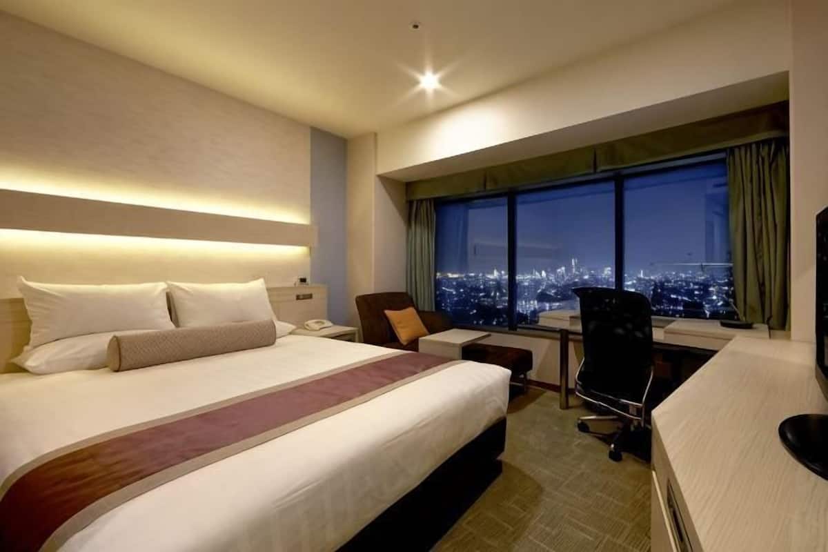 Quarto de hotel com uma cama grande, uma escrivaninha com cadeira, vista para a cidade pela janela e céu noturno visível através da janela.