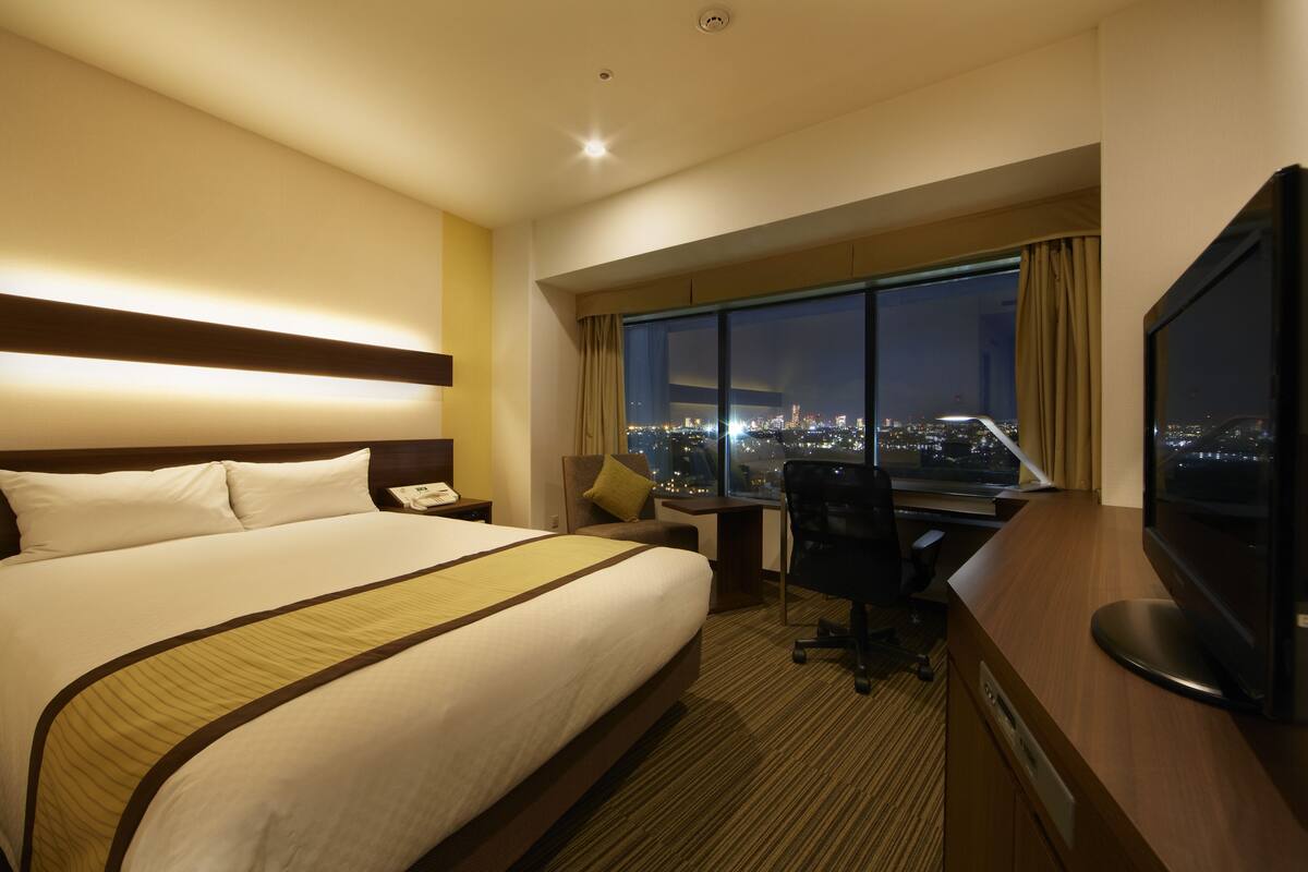 Ein Hotelzimmer mit einem großen Bett, einem Schreibtisch, einem Sessel, einem Fernseher und Blick auf eine beleuchtete Stadt bei Nacht.