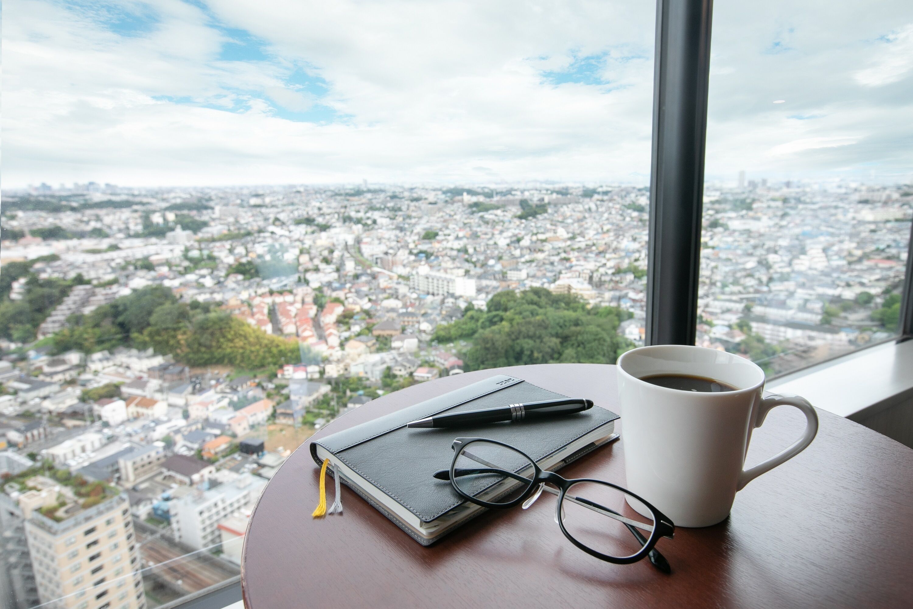 A notebook, glasses, and a coffee cup on a table with a city view through a window.