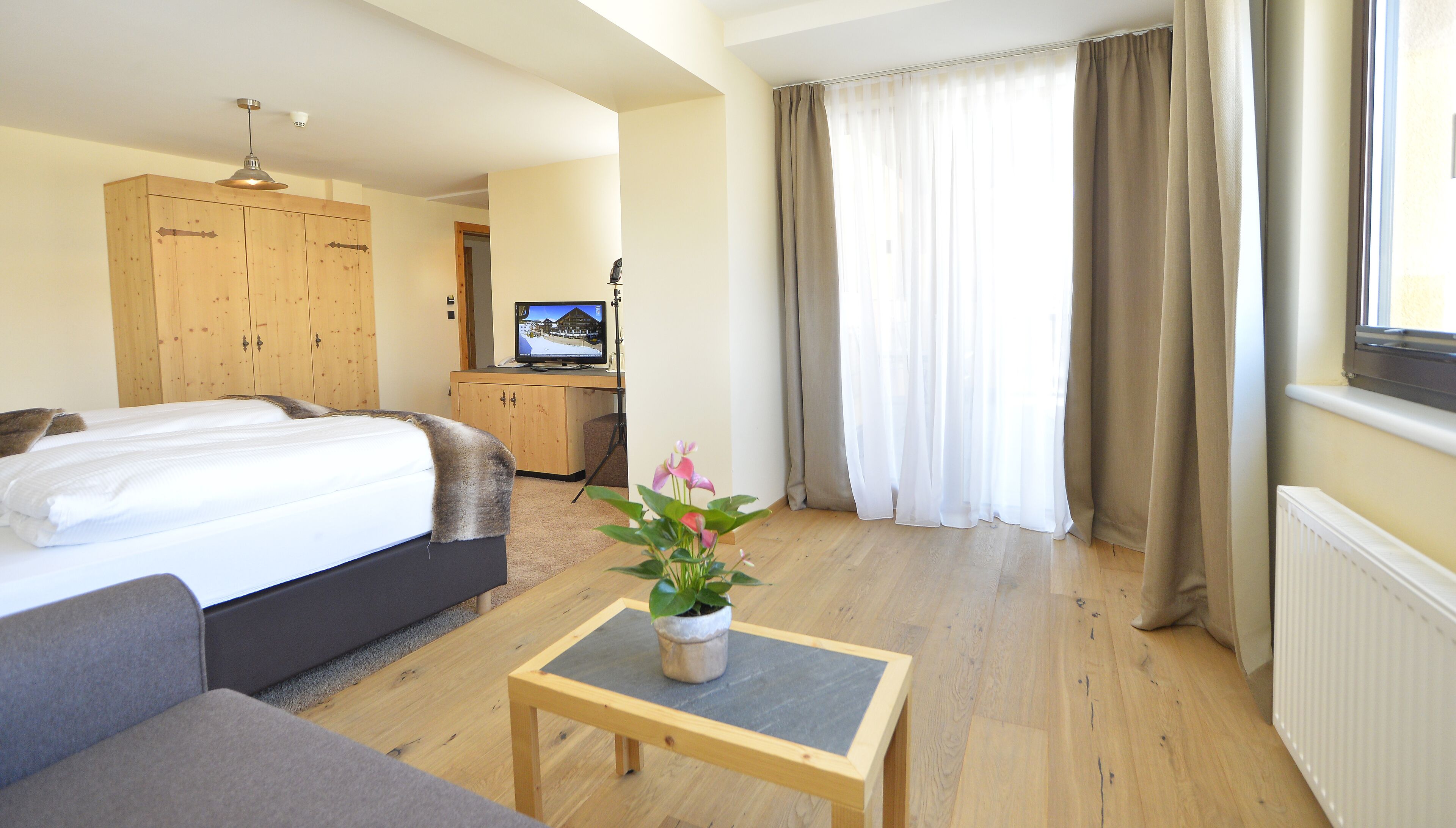A hotel room with a bed, a small table with a plant, a television, and a wooden wardrobe.
