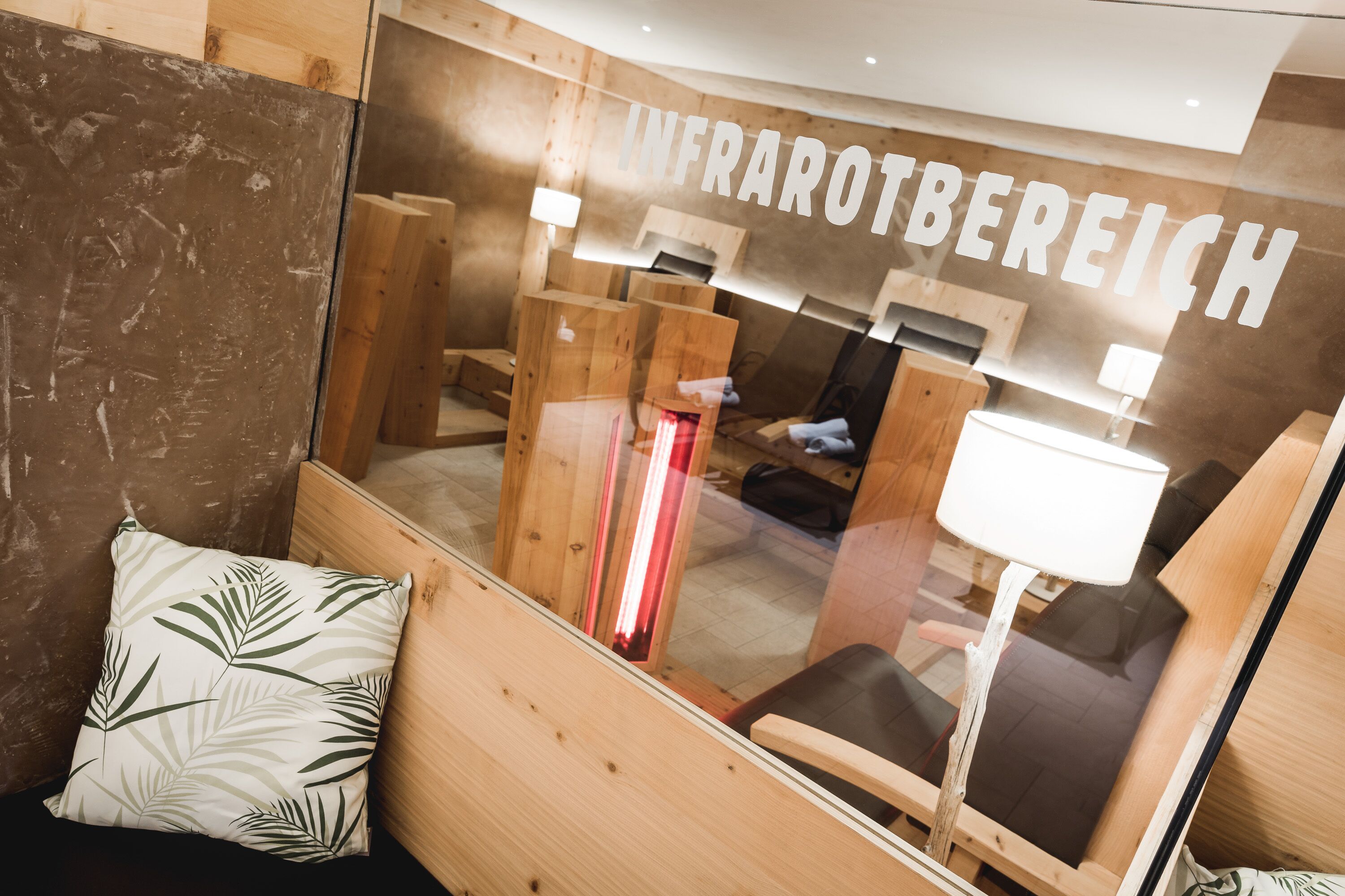 A room with a wooden interior, a glass wall with 'INFRAROTBEREICH' text, a wooden bed, a pillow with a leaf pattern, and a chair with a lamp.