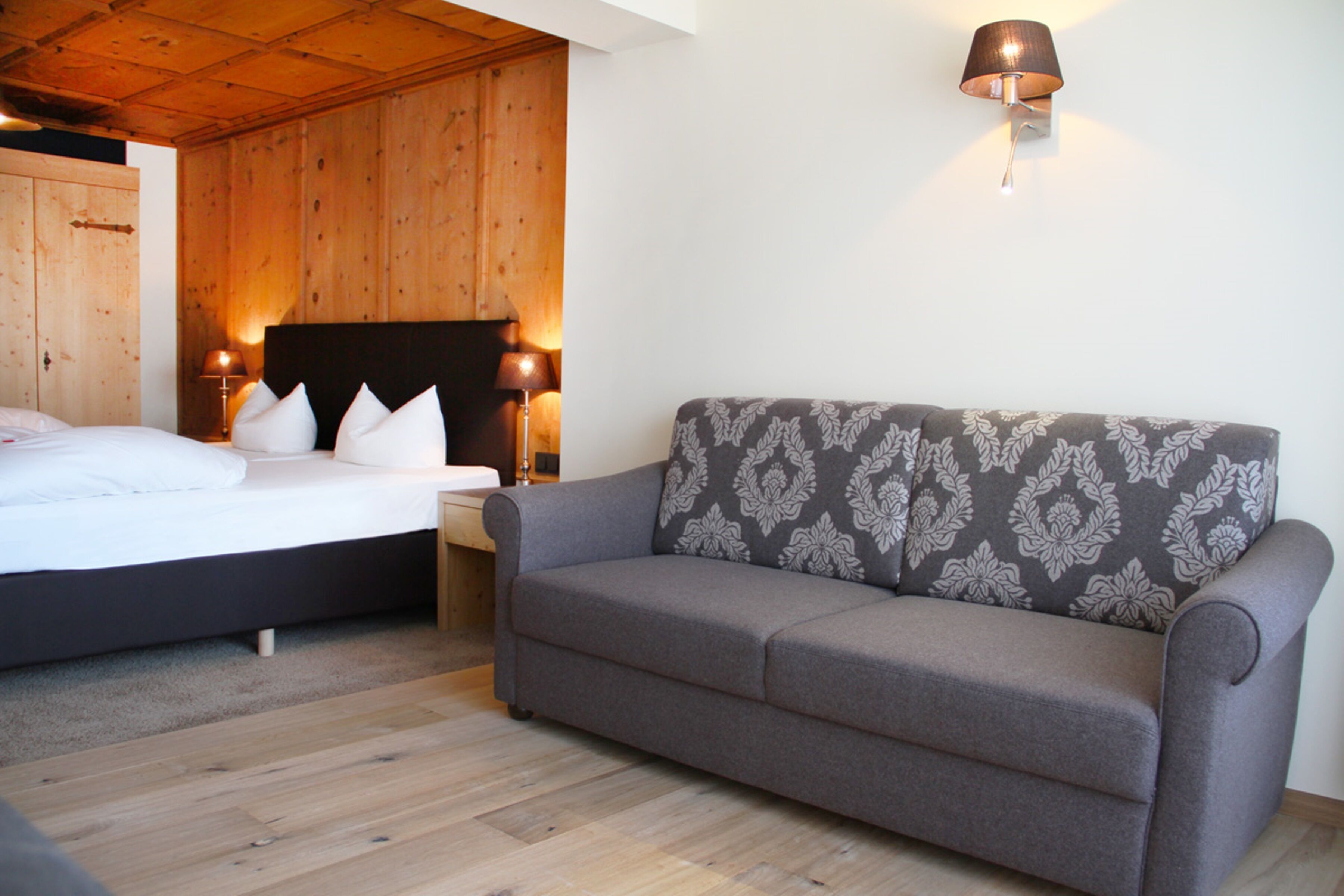 A bedroom with a bed, a grey sofa, wooden walls, and a wall-mounted lamp.