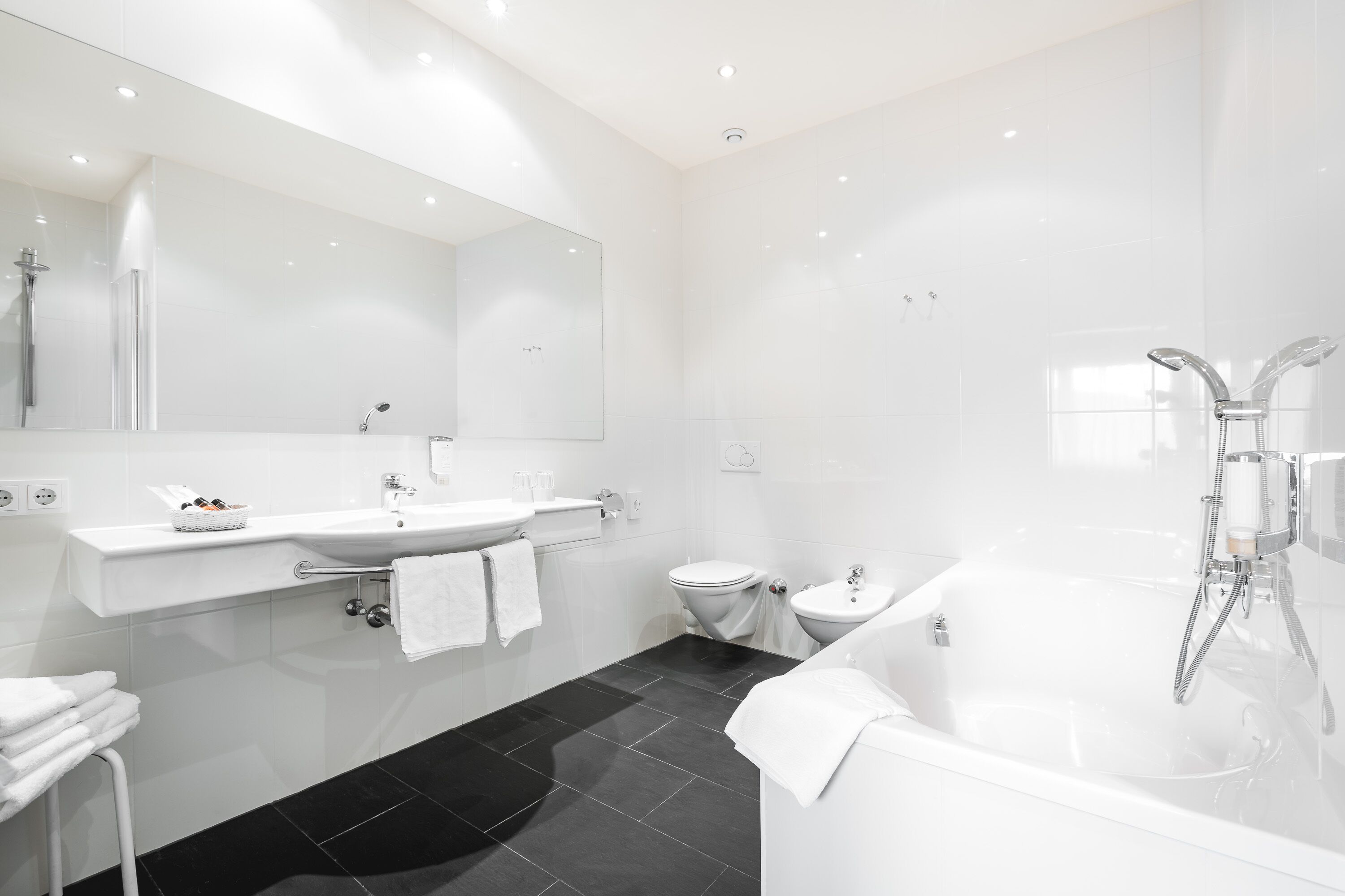 Un baño moderno con un lavamanos blanco, dos inodoros, una bañera y un espejo grande.