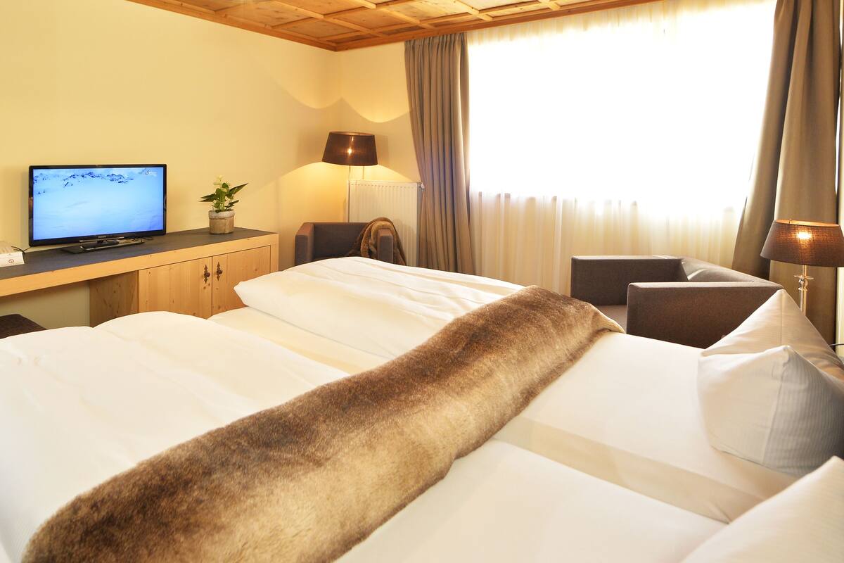 A hotel room with a bed, a desk with a computer, a chair, a lamp, and a window with curtains.