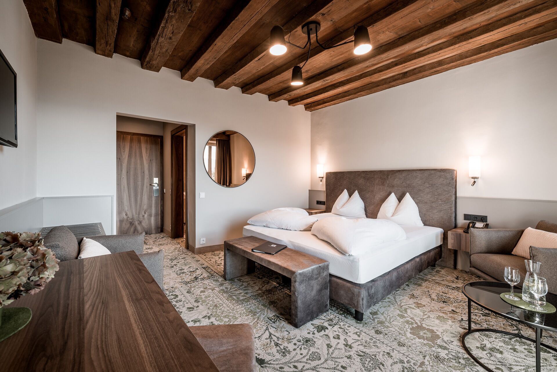 Una habitación de hotel moderna con una cama grande, techo de madera, espejo redondo y un pequeño rincón de estar con mesa y sillas.