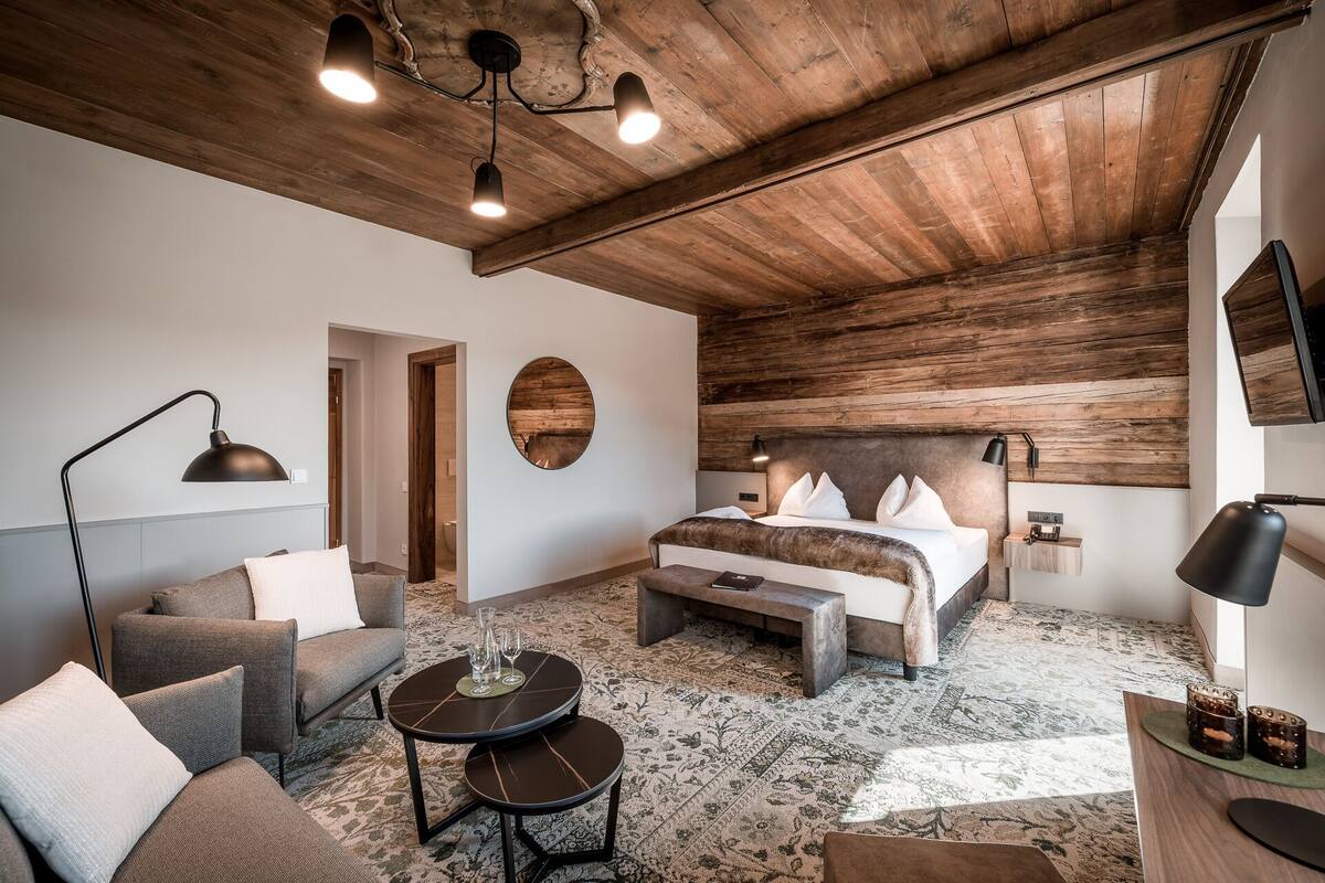 A modern bedroom with a wooden ceiling, a large bed with a headboard, a sofa, a coffee table, and a TV mounted on the wall.
