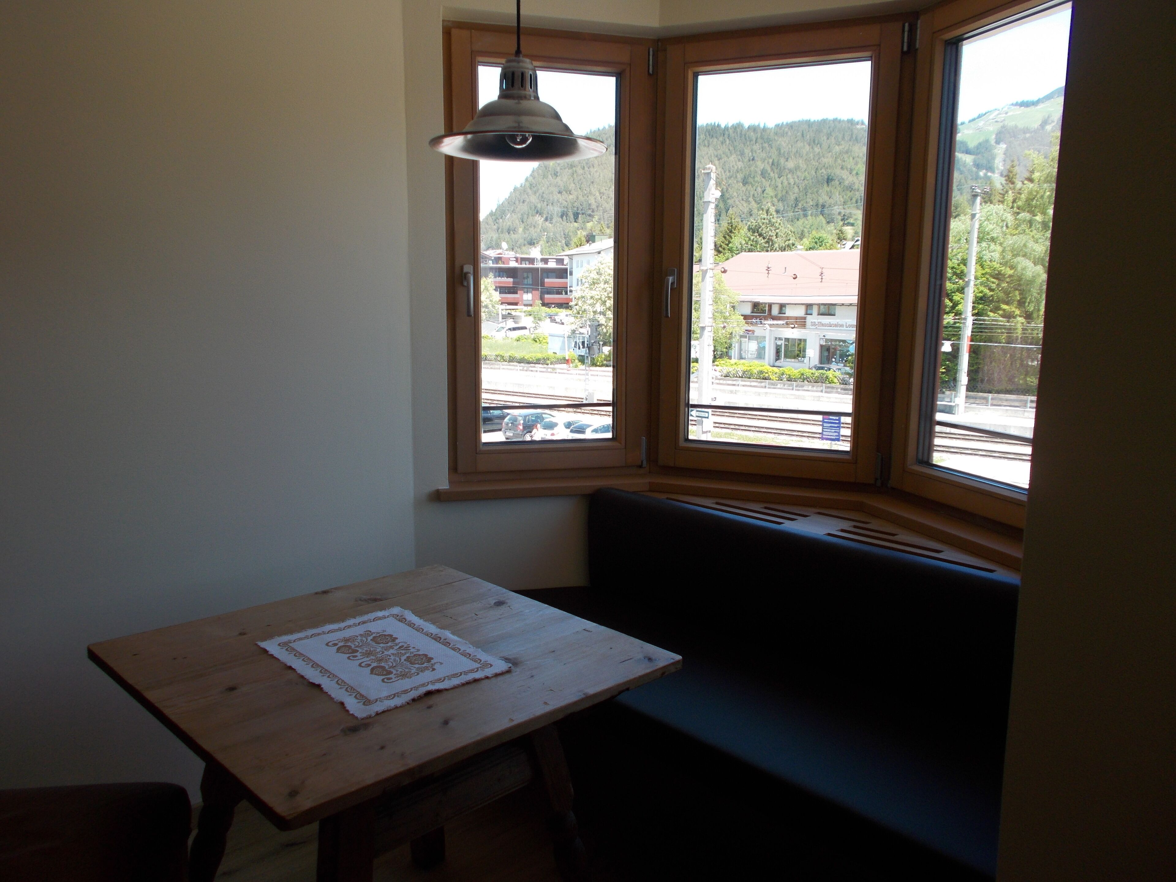 A corner room with a wooden table, a bench, and a window with a view of a forested hill and buildings.
