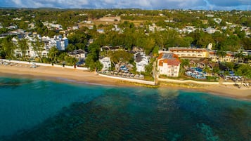 Vista aérea de uma cidade costeira com praia, águas azuis cristalinas e diversos edifícios, incluindo um hotel com piscina e guarda-sóis.