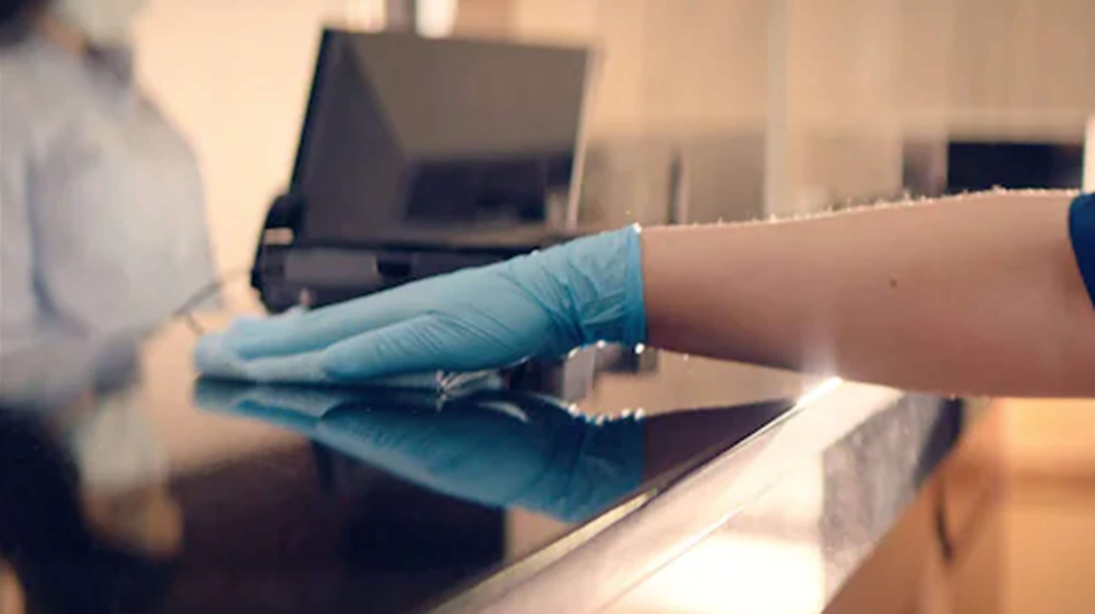 Une main gantée de bleu essuie une surface réfléchissante, probablement un comptoir ou une table.