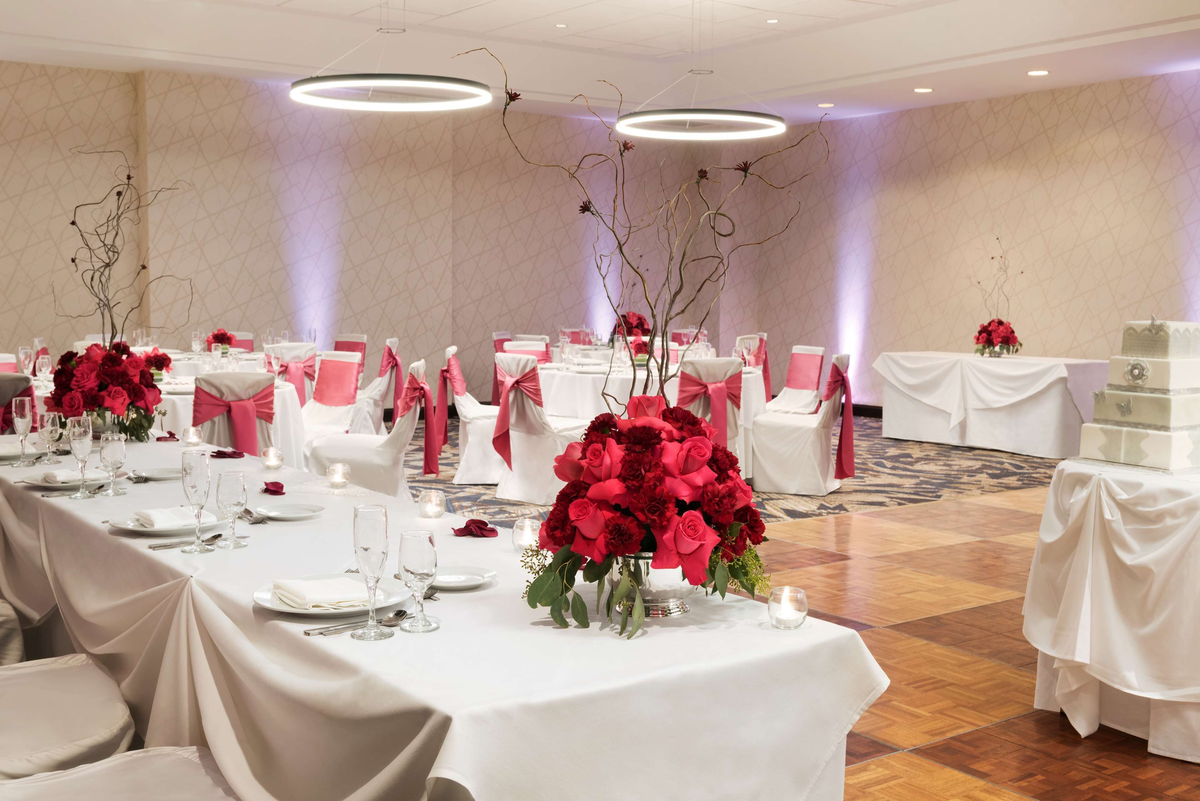 A banquet hall with tables set for a formal event, featuring white tablecloths, pink chair covers, and red floral centerpieces.