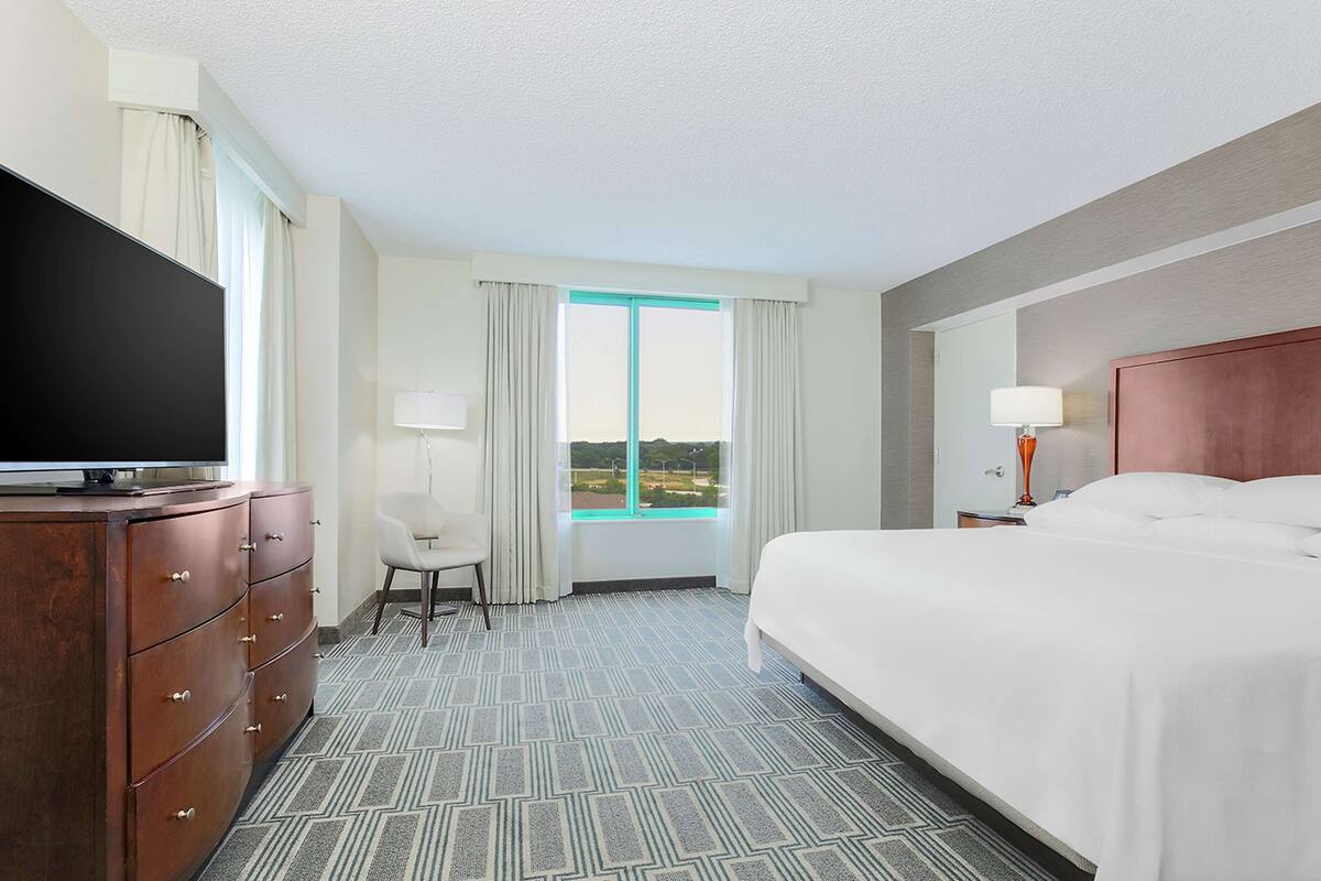 Une chambre d’hôtel avec un grand lit, un téléviseur posé sur une commode en bois, une chaise et une fenêtre avec des rideaux.