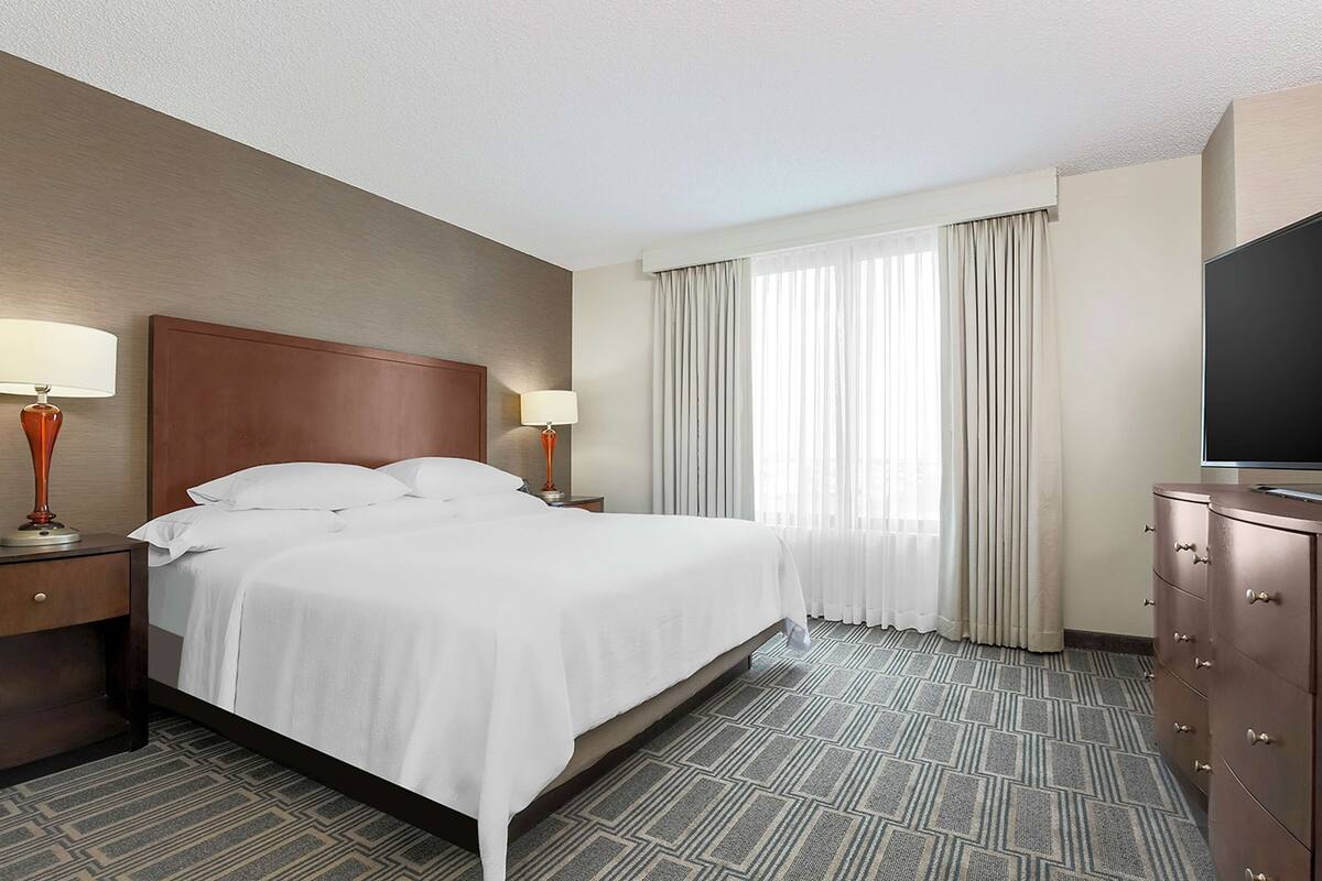 Une chambre d’hôtel avec un grand lit, deux tables de chevet avec des lampes, une télévision et une fenêtre avec des rideaux.