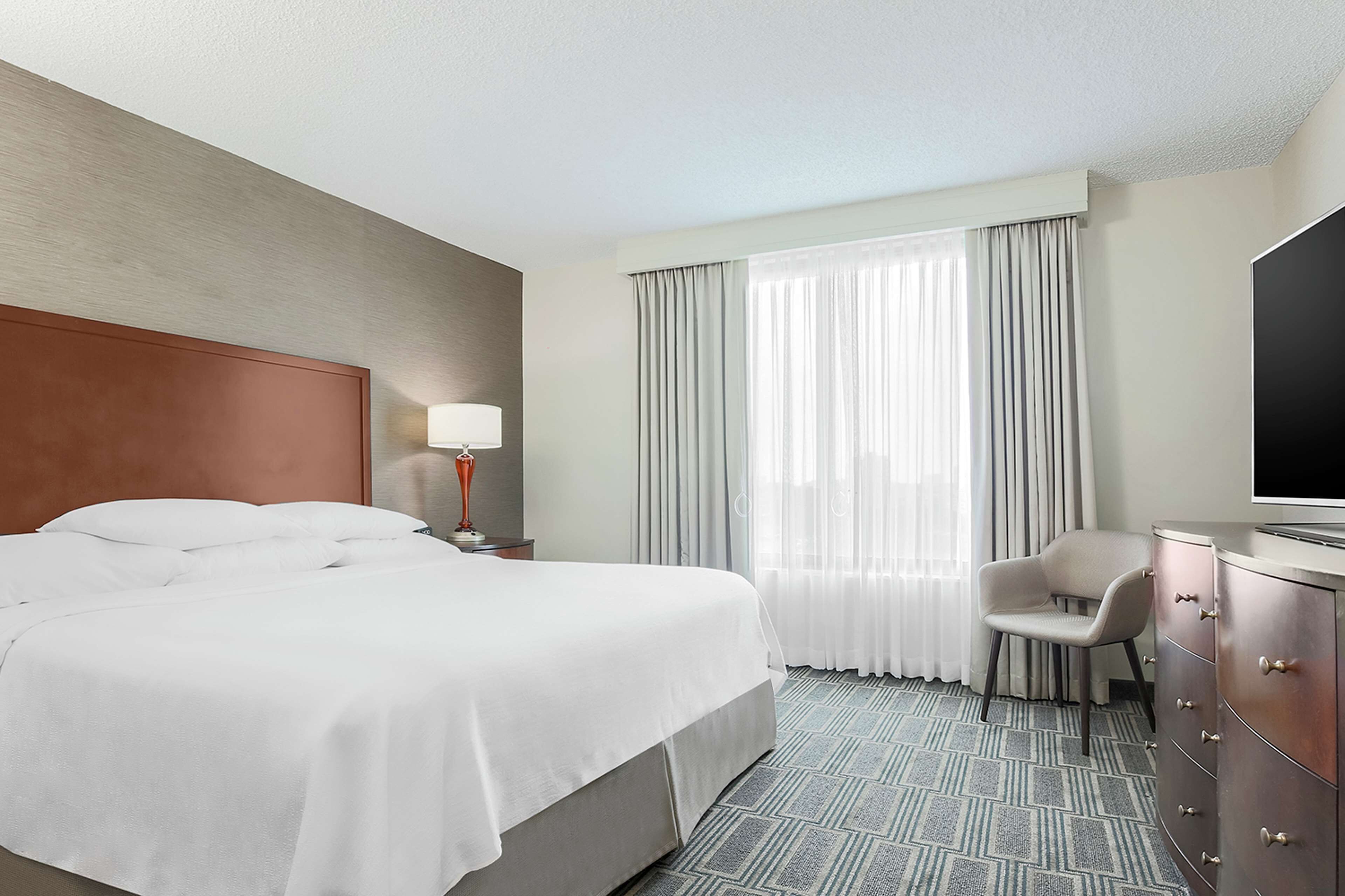 A hotel room with a large bed, a desk with a flat-screen TV, a chair, and a window with curtains.