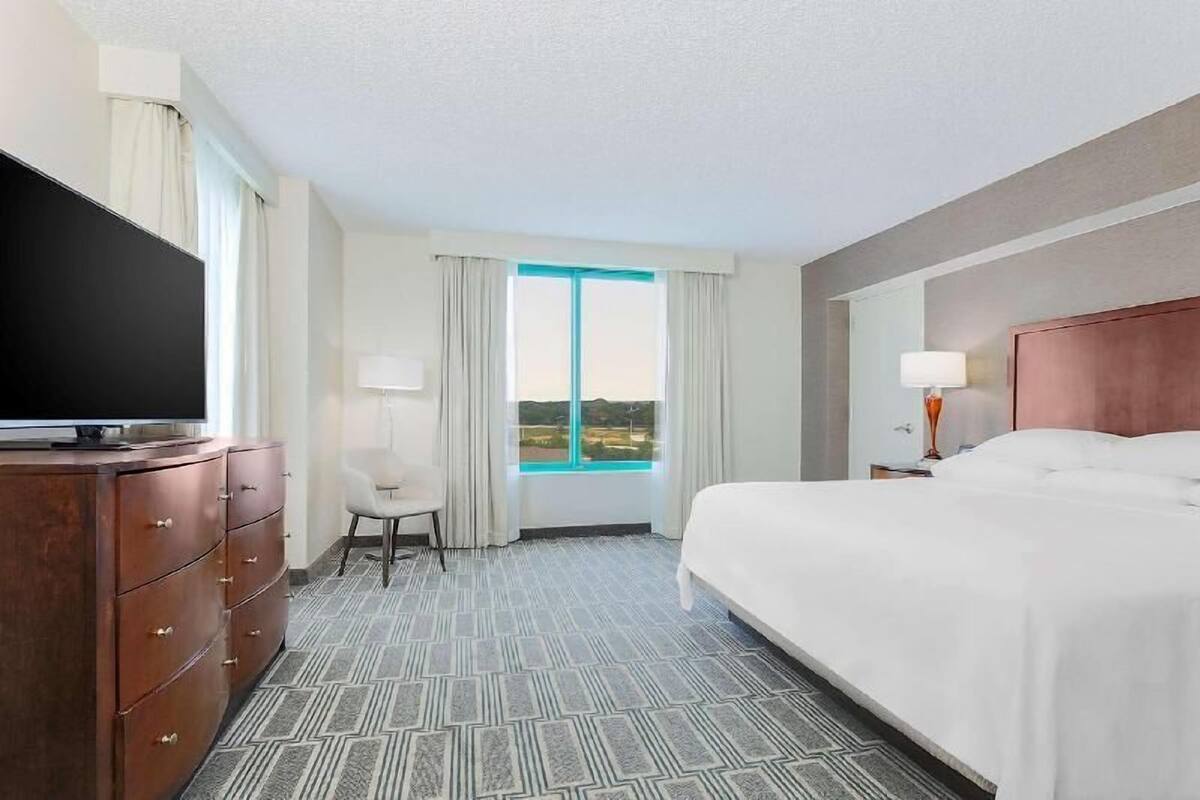 Une chambre d’hôtel avec un lit, une télévision, un bureau, une chaise et une fenêtre avec des rideaux.
