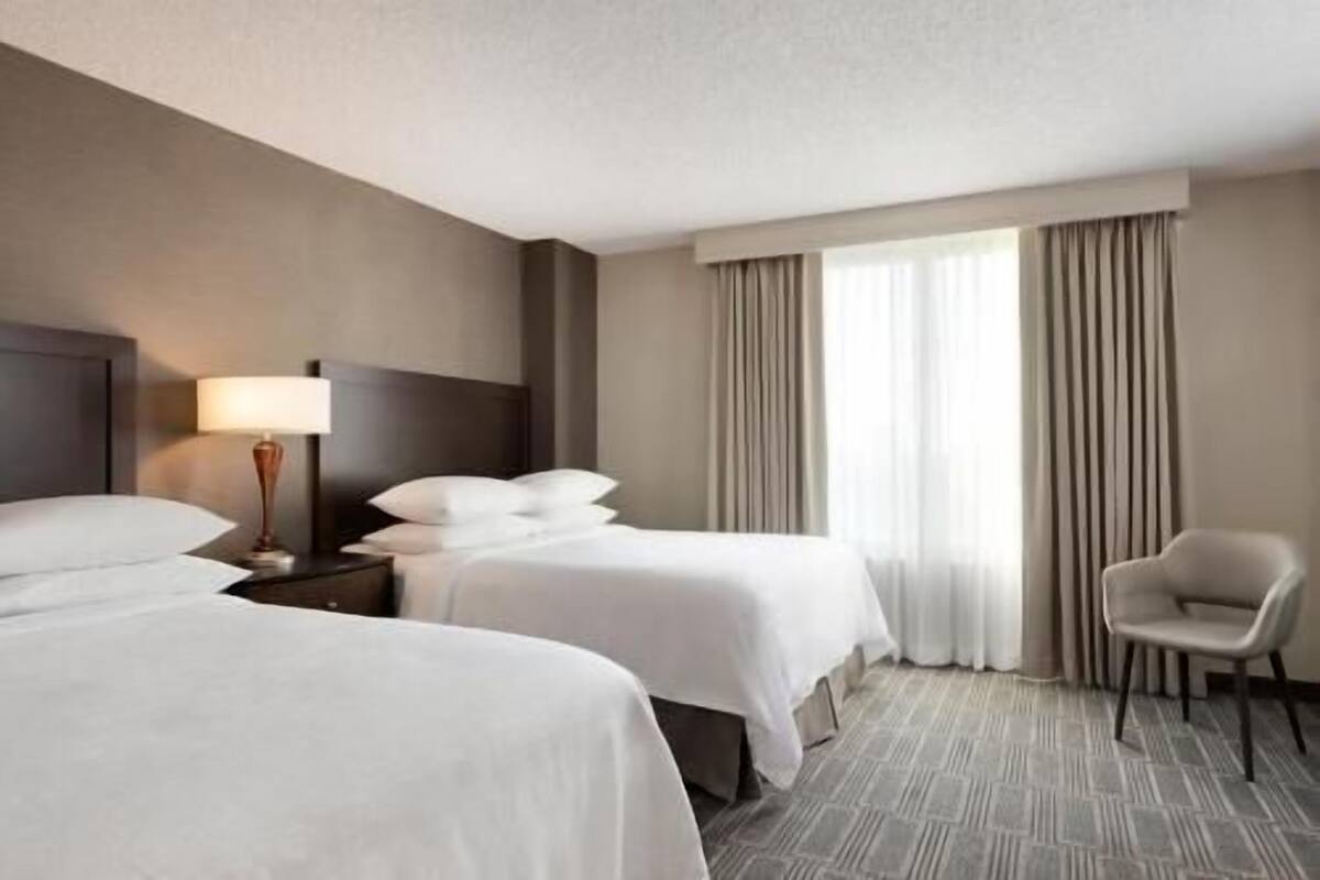 Une chambre d’hôtel avec deux lits, une chaise, une lampe et une grande fenêtre avec des rideaux.