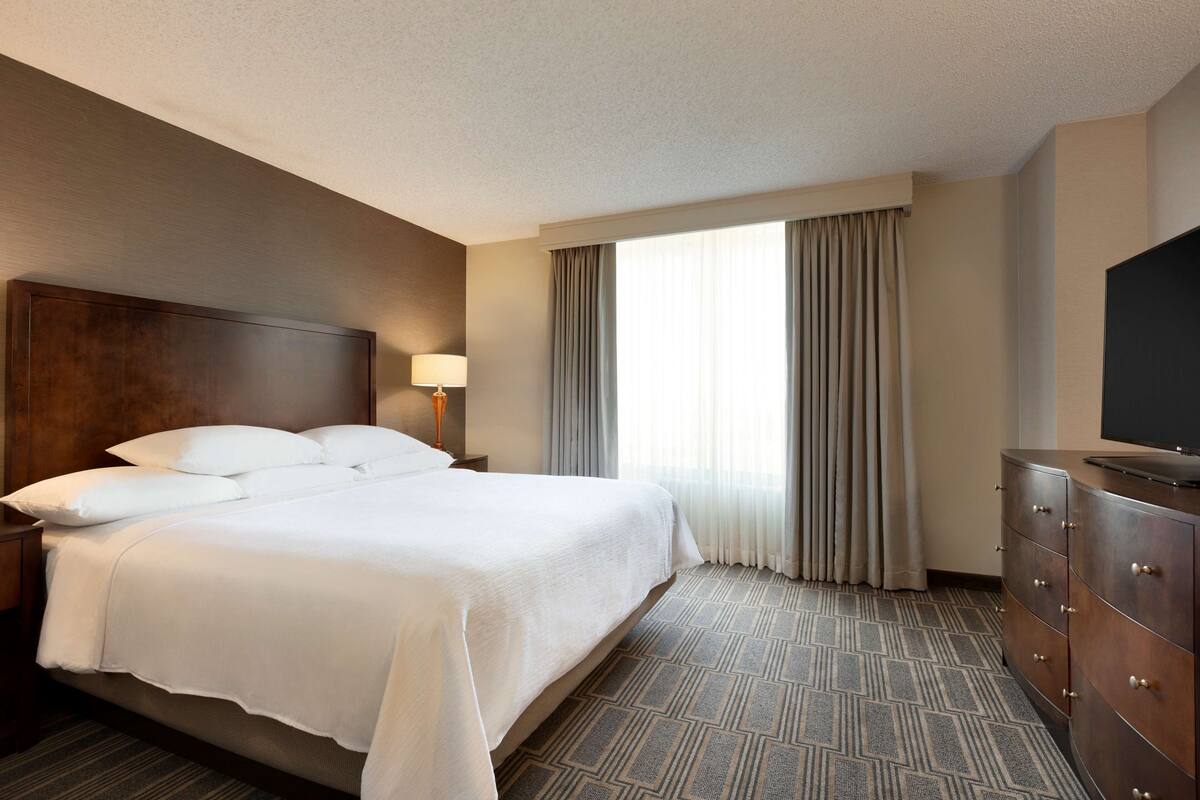 Une chambre d’hôtel avec un grand lit, une commode, une télévision et une fenêtre avec des rideaux.