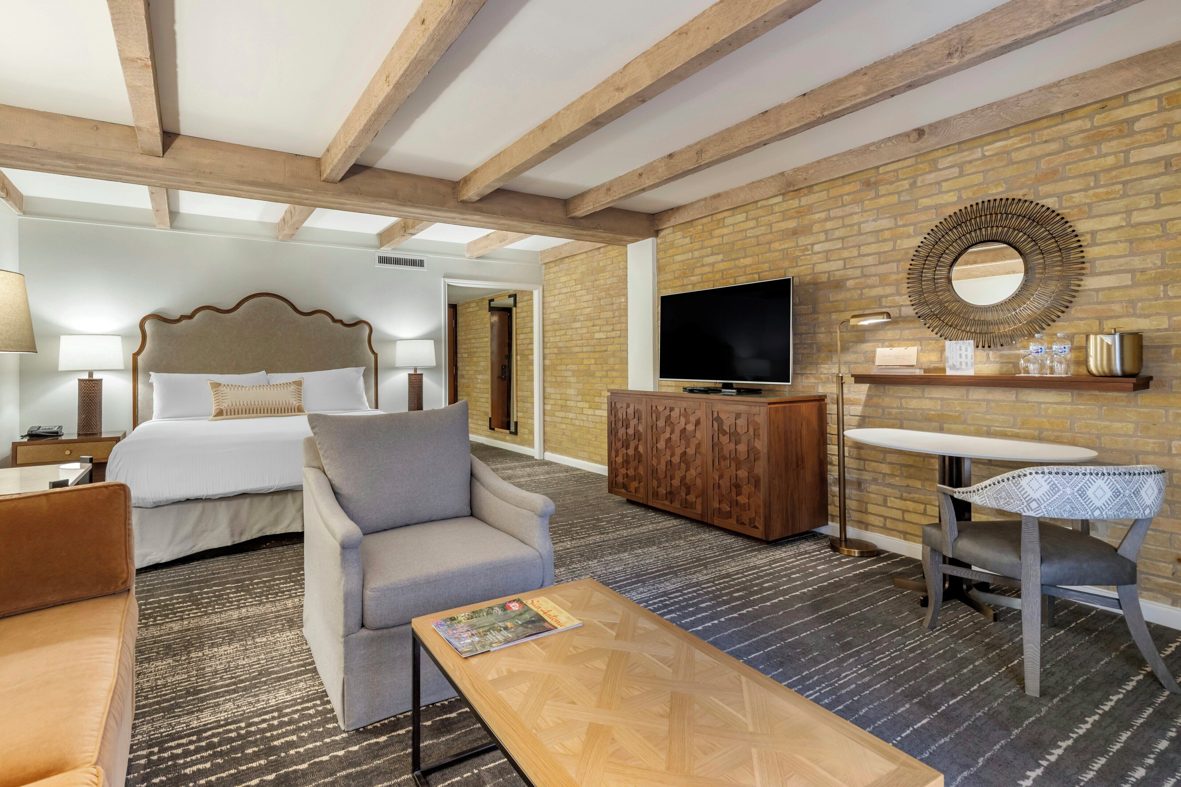 Habitación de hotel con cama, sofá, mesa de centro, televisor, espejo y pared de ladrillo.