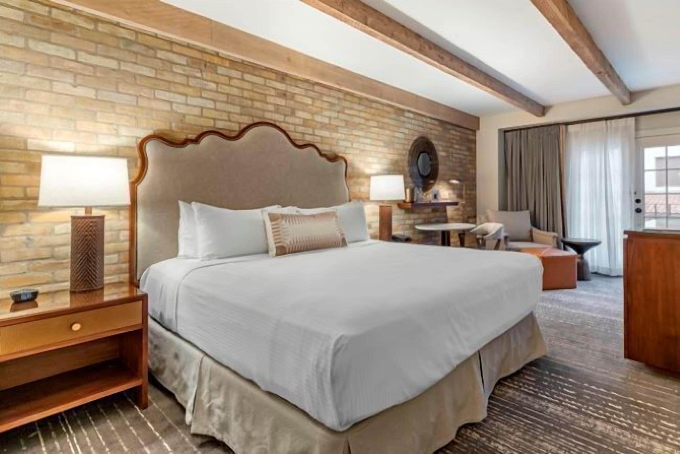 Habitación de hotel con una cama grande, cabecera de madera, pared de ladrillo y una mesita con lámpara.