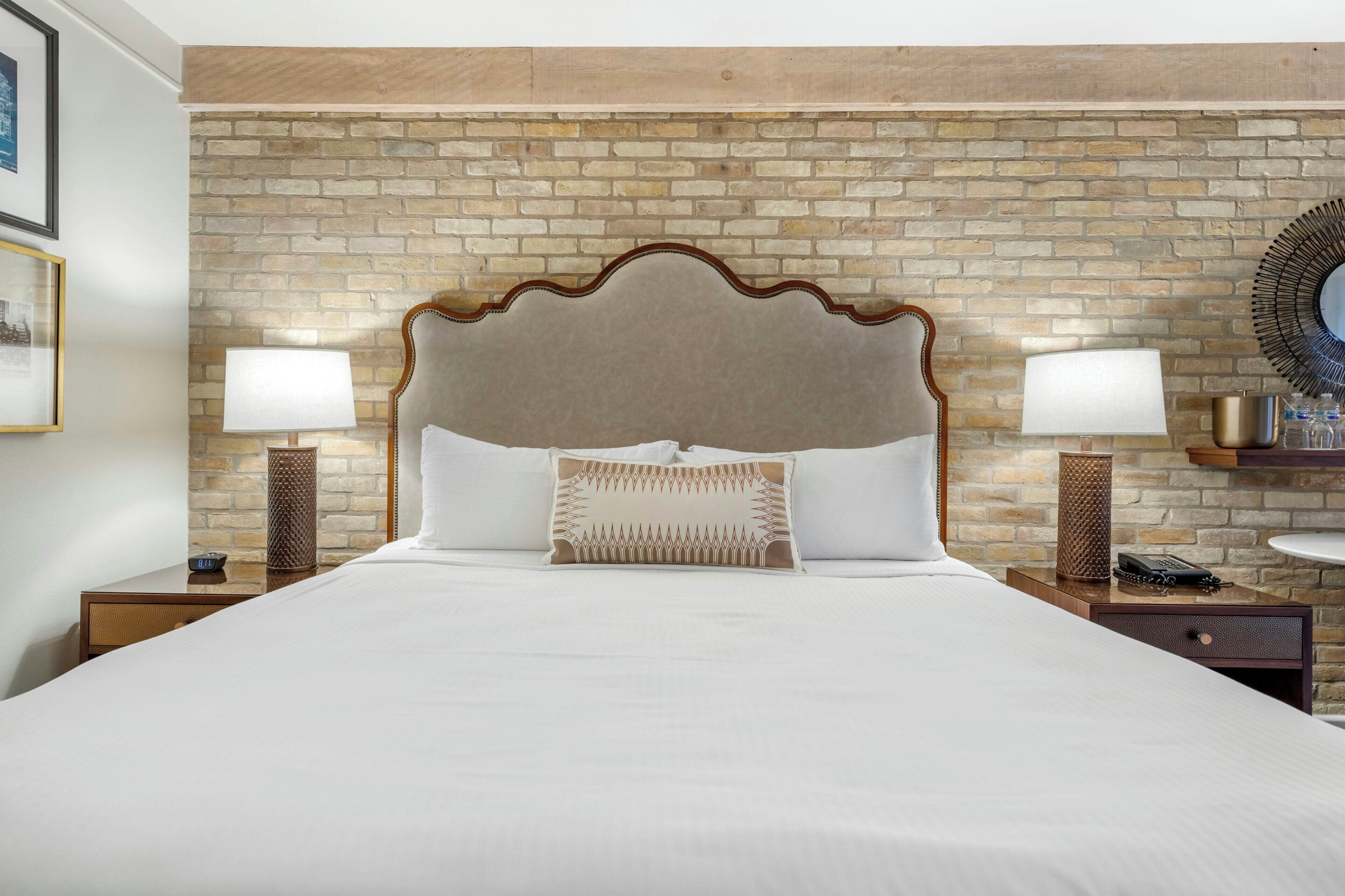 A hotel room with a brick-patterned wall, a large bed with a wooden headboard, two bedside tables with lamps, and a round mirror on the wall.