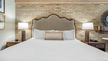 A hotel room with a brick-patterned wall, a large bed with a wooden headboard, two bedside tables with lamps, and a round mirror on the wall.