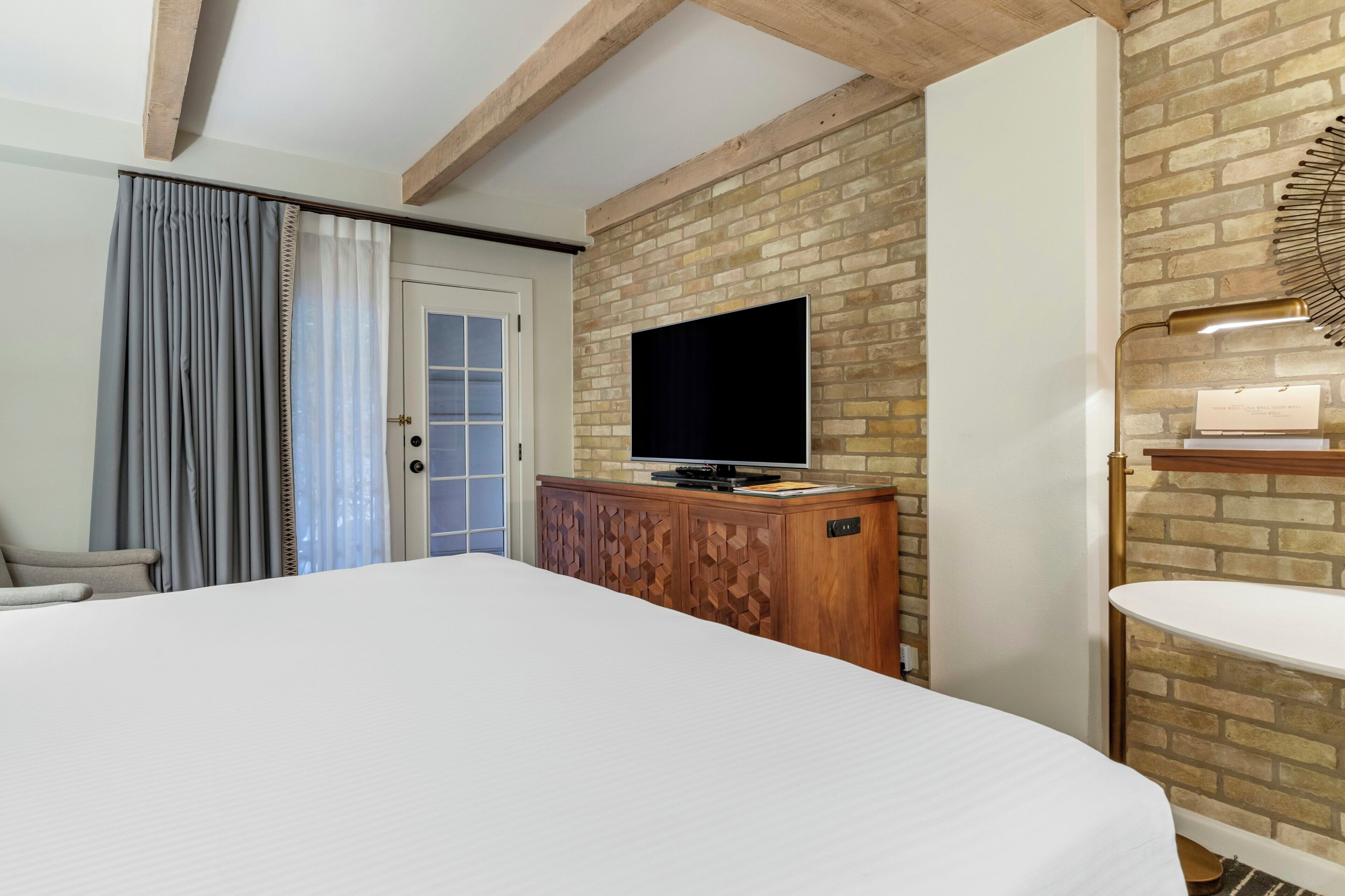 Una camera da letto con un letto grande, una televisione montata su una parete in mattoni, un armadietto in legno e un tavolino con una lampada.