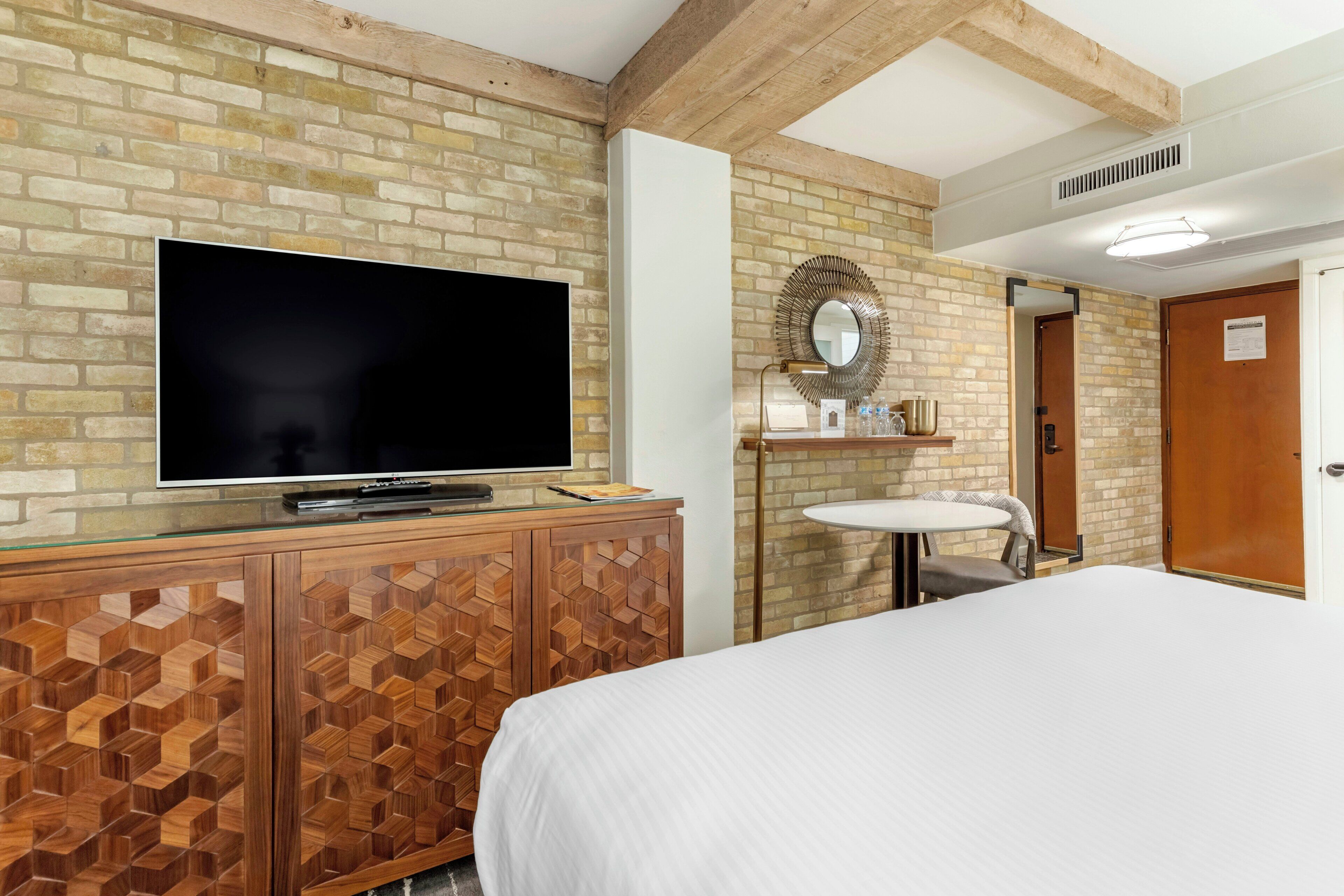 A hotel room with a brick wall, a flat-screen TV on a wooden cabinet, a round mirror above a small sink, and a bed with white bedding.