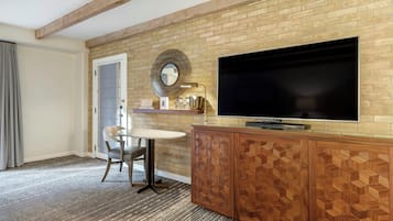 Quarto de hotel com parede de tijolos, uma televisão de tela plana presa a um armário de madeira, uma mesa pequena com uma cadeira e uma porta com cortina estampada.