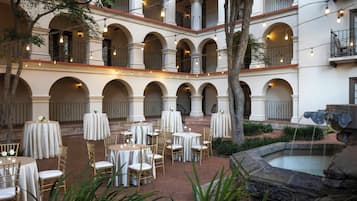 Un patio con varias mesas y sillas, rodeado de arcos y columnas, y una fuente.
