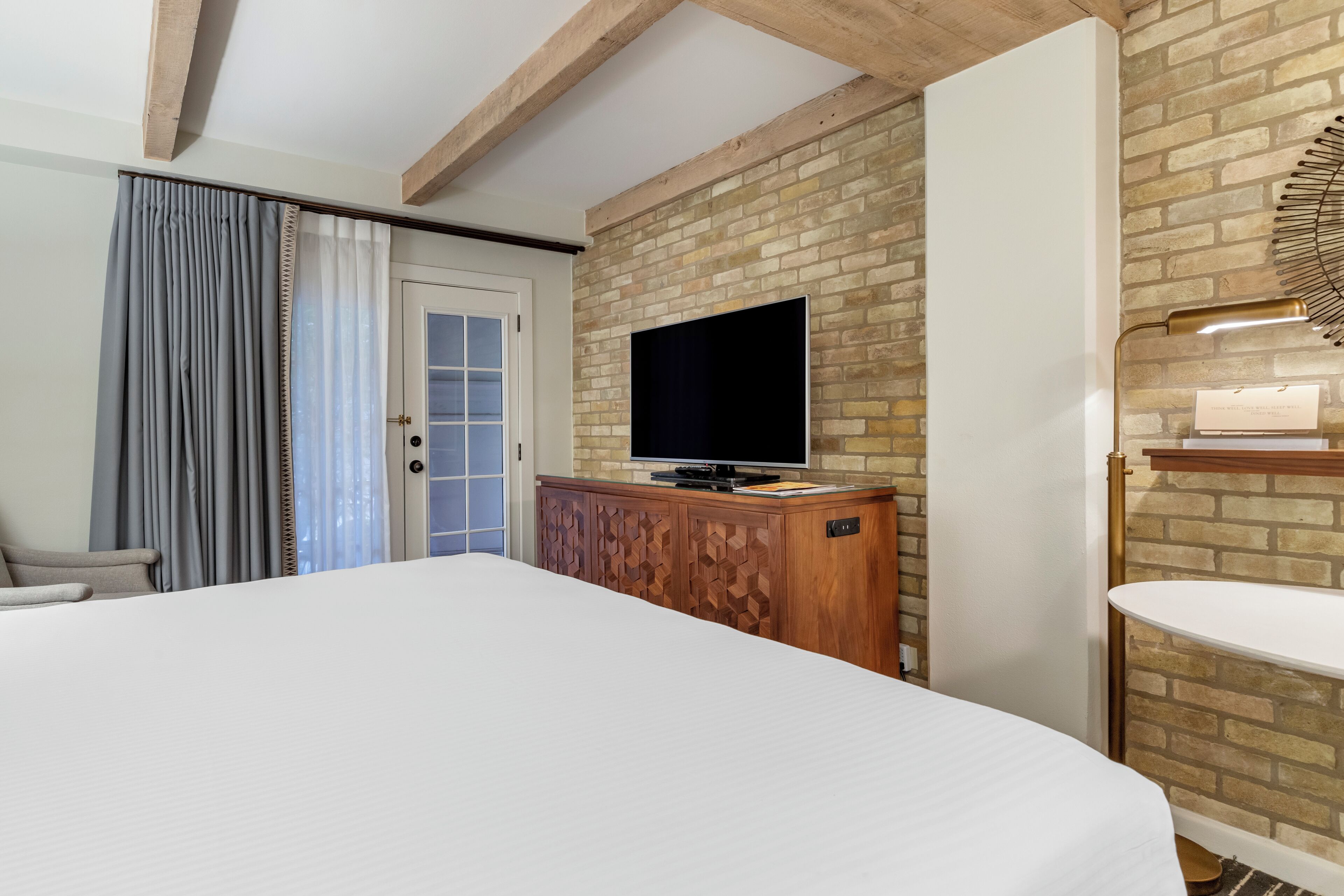 Habitación de hotel con una cama grande, un televisor montado en una pared de ladrillo, un armario de madera y una mesita de noche con una lámpara.