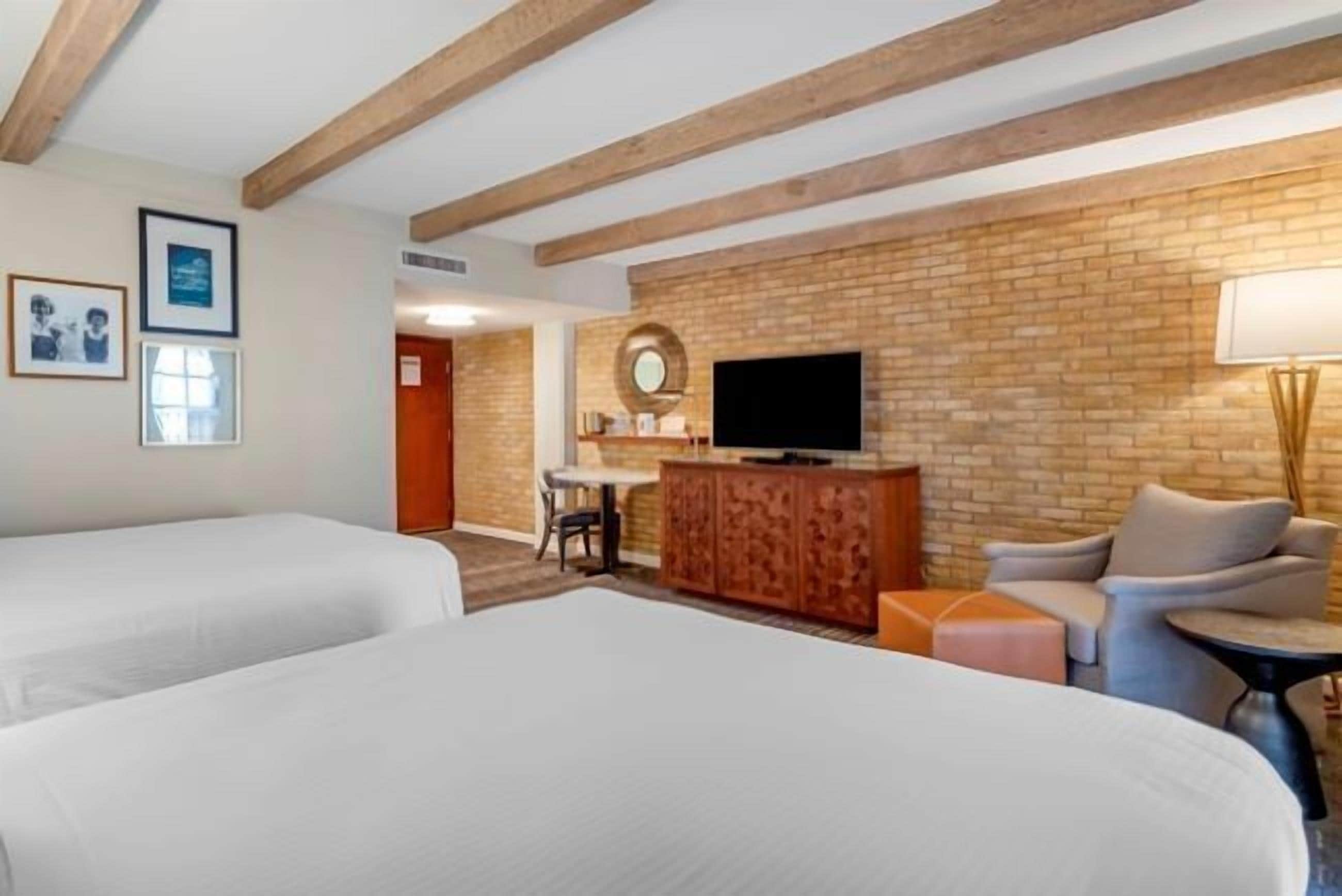 Quarto de hotel com duas camas, uma televisão, um armário de madeira e uma parede de tijolos.