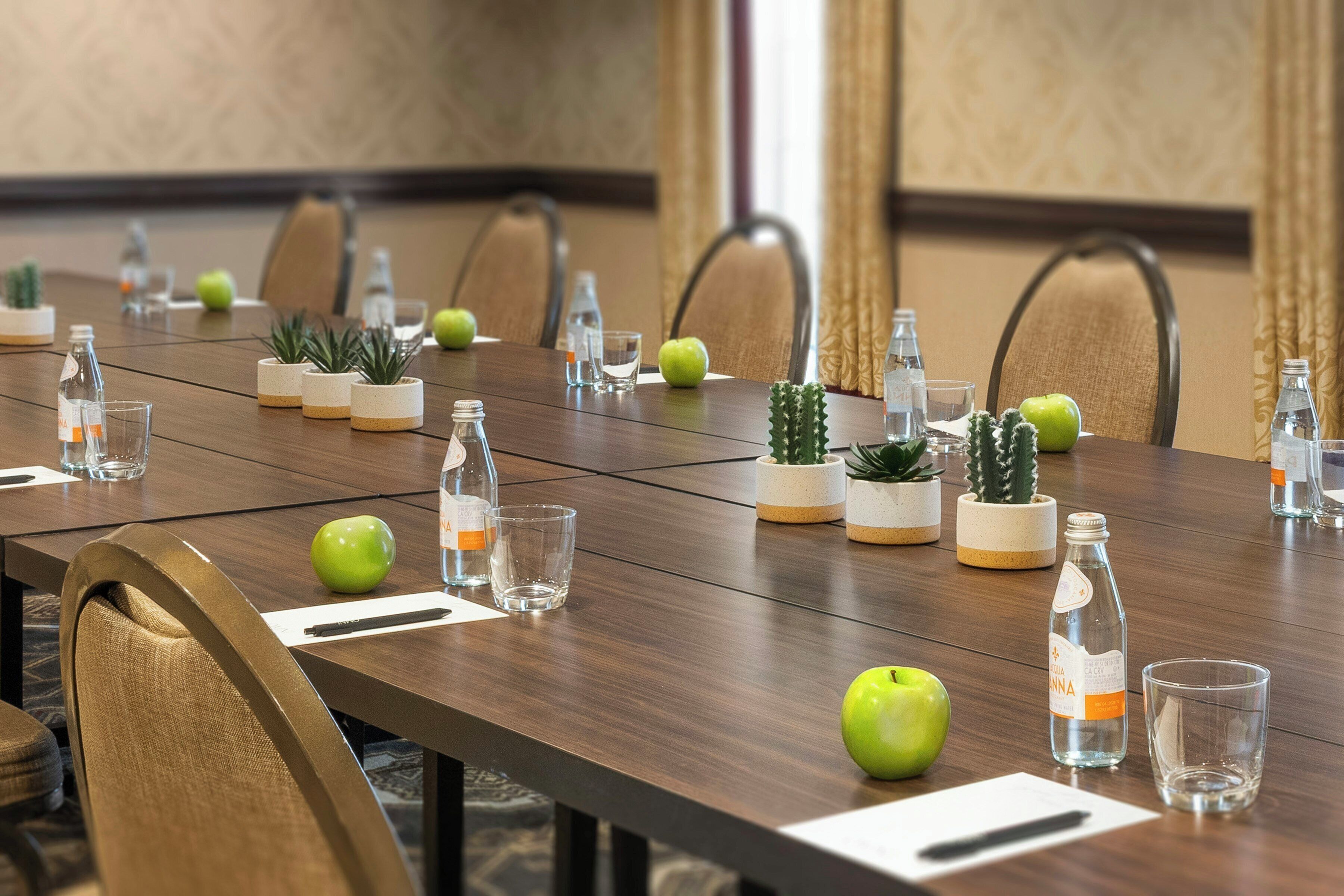 Un salón de conferencias con una larga mesa, sillas, botellas de agua, vasos y pequeñas plantas en macetas.