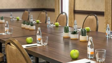 Un salón de conferencias con una larga mesa, sillas, botellas de agua, vasos y pequeñas plantas en macetas.