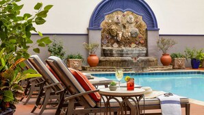 A poolside area with lounge chairs, a table with a drink, and a decorative wall with a fountain. - Omni La Mansion del Rio (San Antonio)