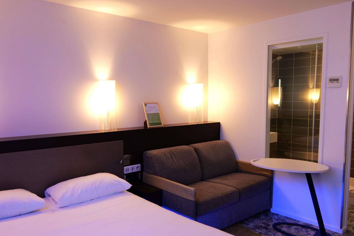 A hotel room with a bed, a sofa, a small table, and two wall-mounted lights.