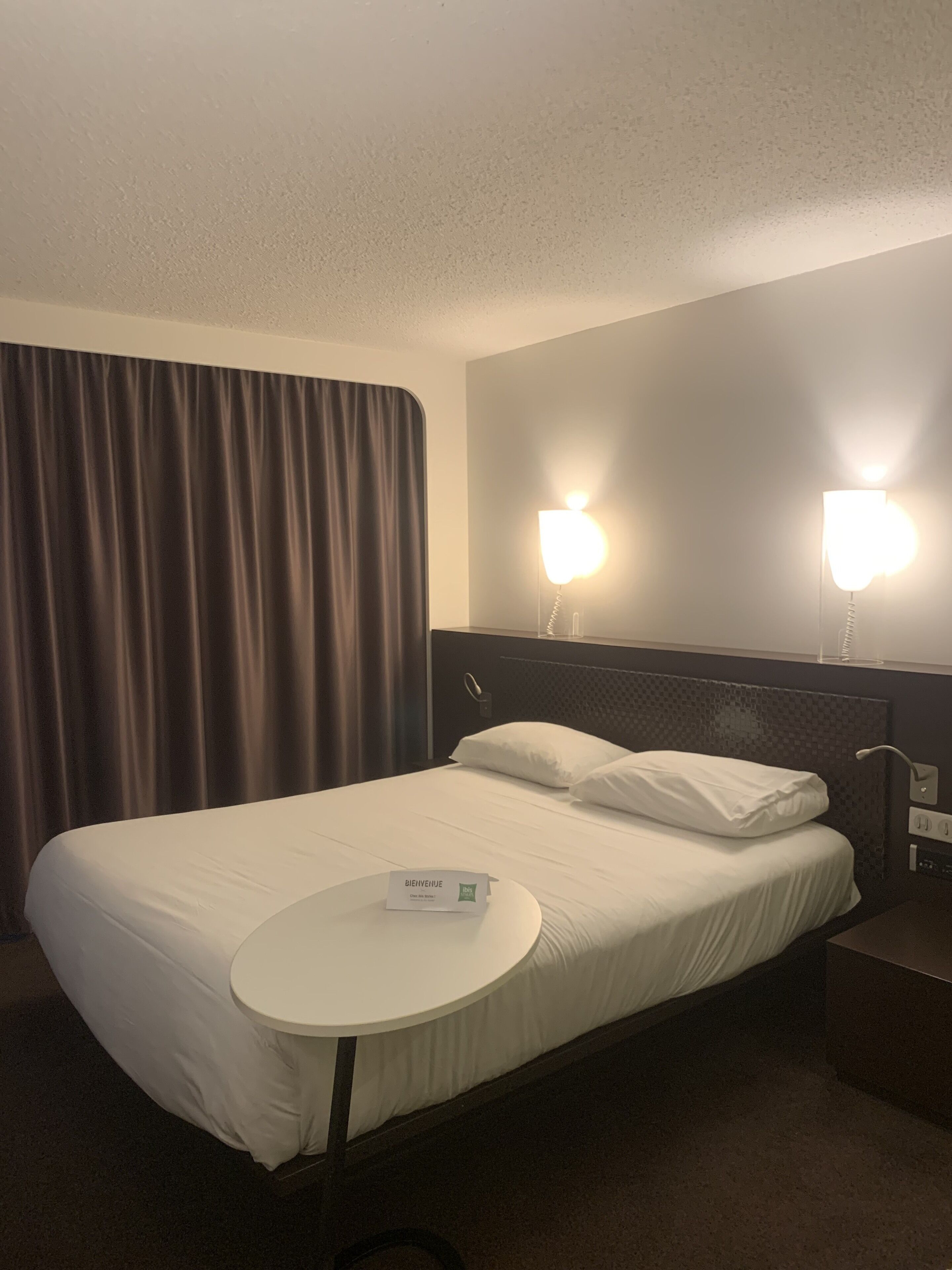Habitació d'hotel amb un llit ben arropat, una taula de nit amb un cartell de benvinguda i dues làmpades penjades a la paret.