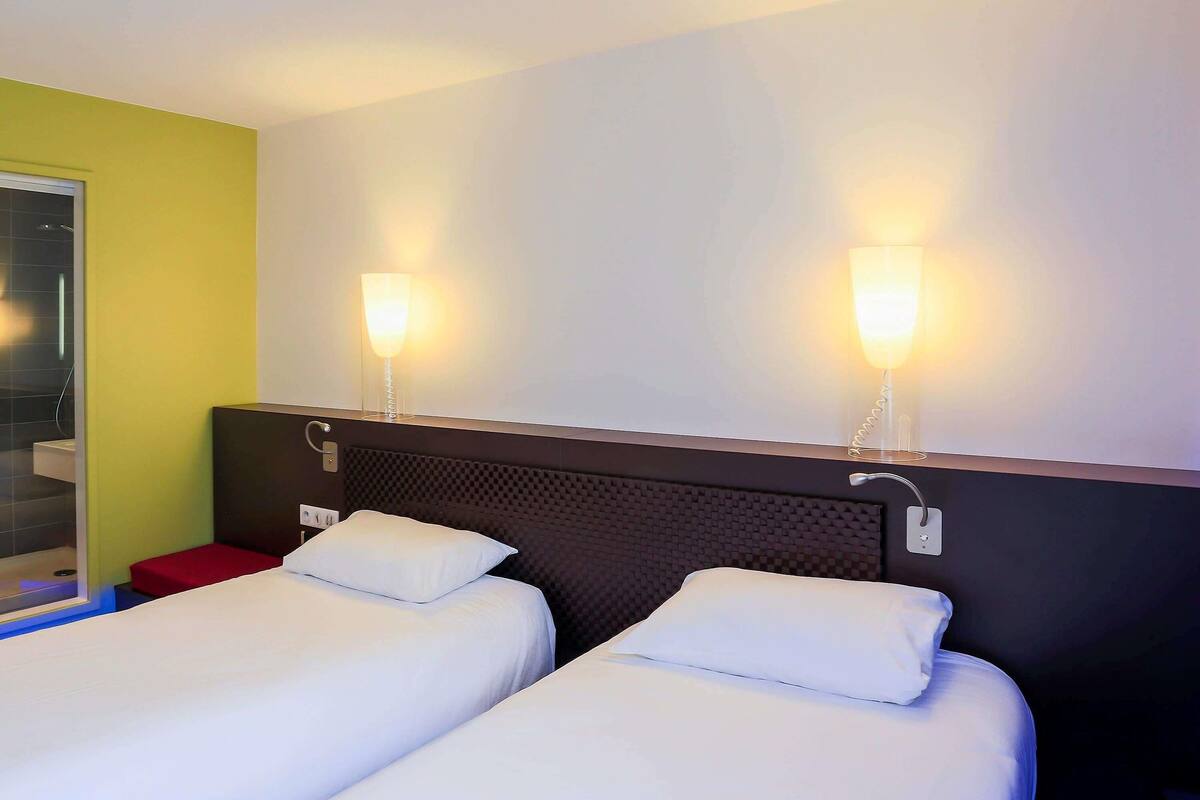 A hotel room with two beds, a red nightstand, a mirror, and wall-mounted lights.