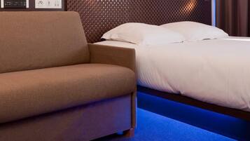 Una habitación de hotel moderna con sofá, cama con ropa blanca, cabecero de madera y alfombra azul.