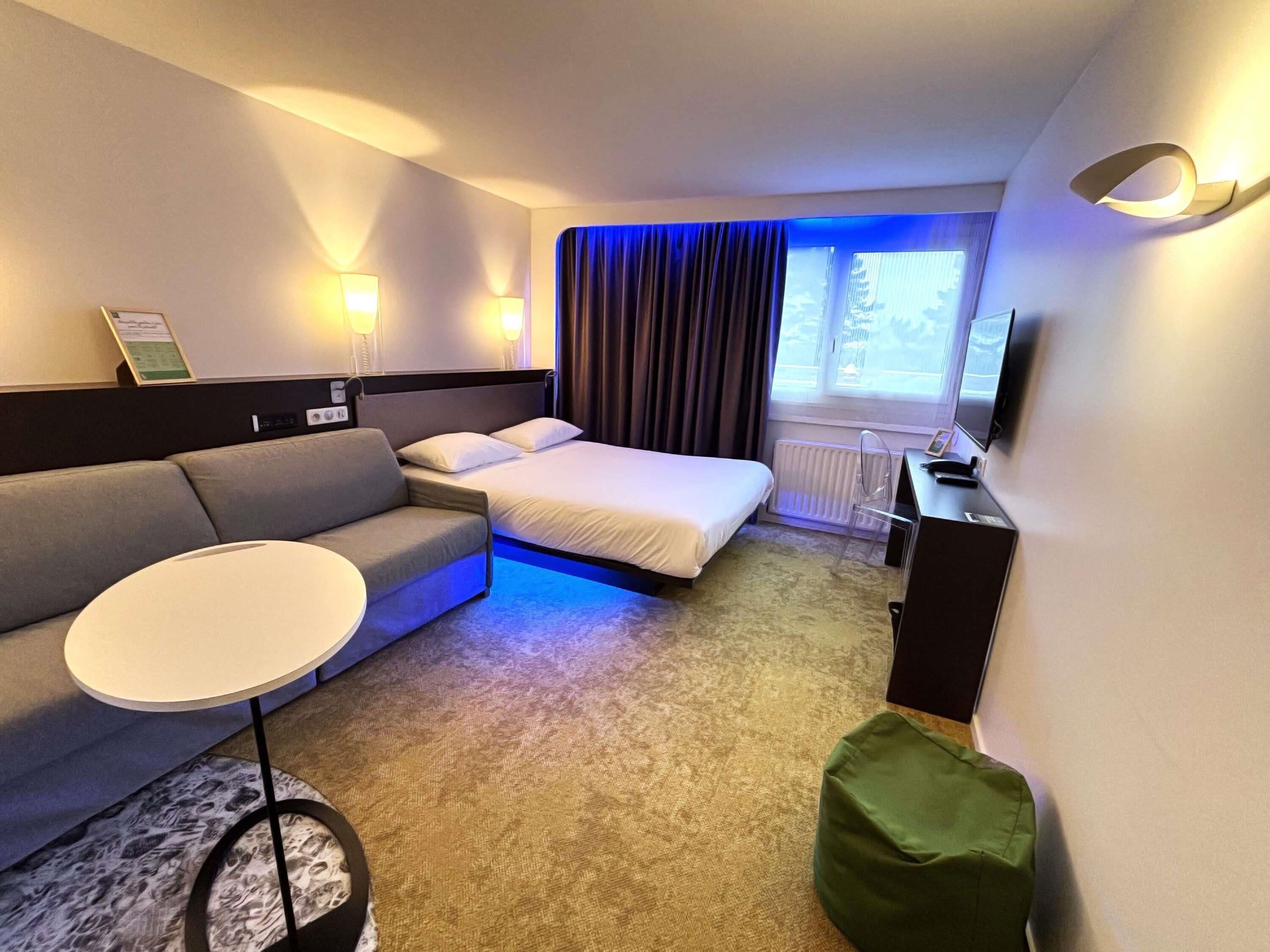 Habitación de hotel con cama, sofá, una mesita, un reposapiés verde, escritorio con silla, televisor y ventana con cortinas.