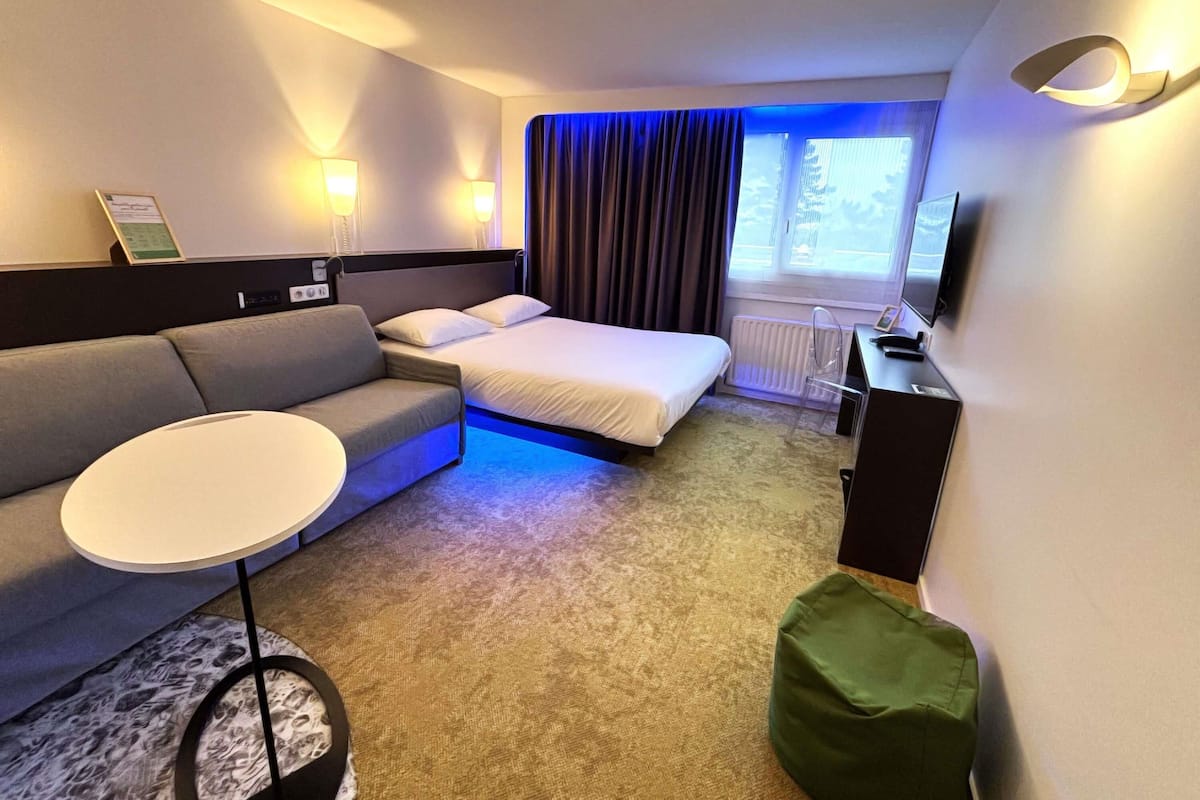 Habitación de hotel con cama, sofá, una mesita, un reposapiés verde, escritorio con silla, televisor y ventana con cortinas.