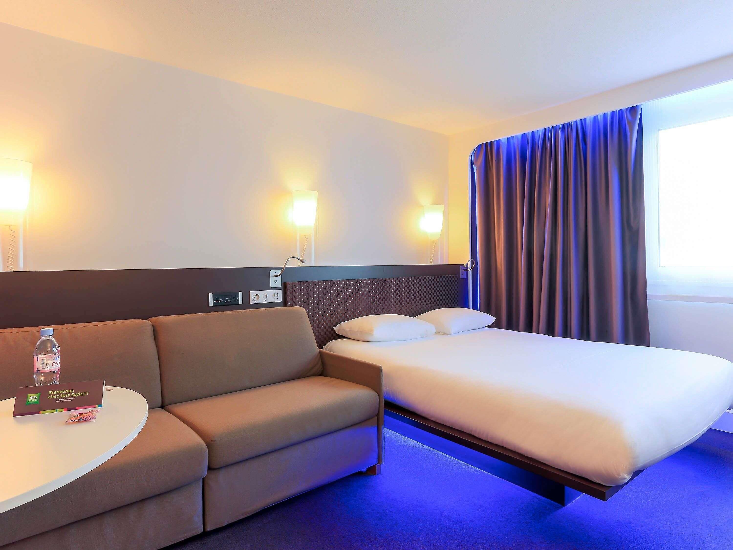 Habitación de hotel con cama, sofá, una mesita con una botella y teléfono en la pared.