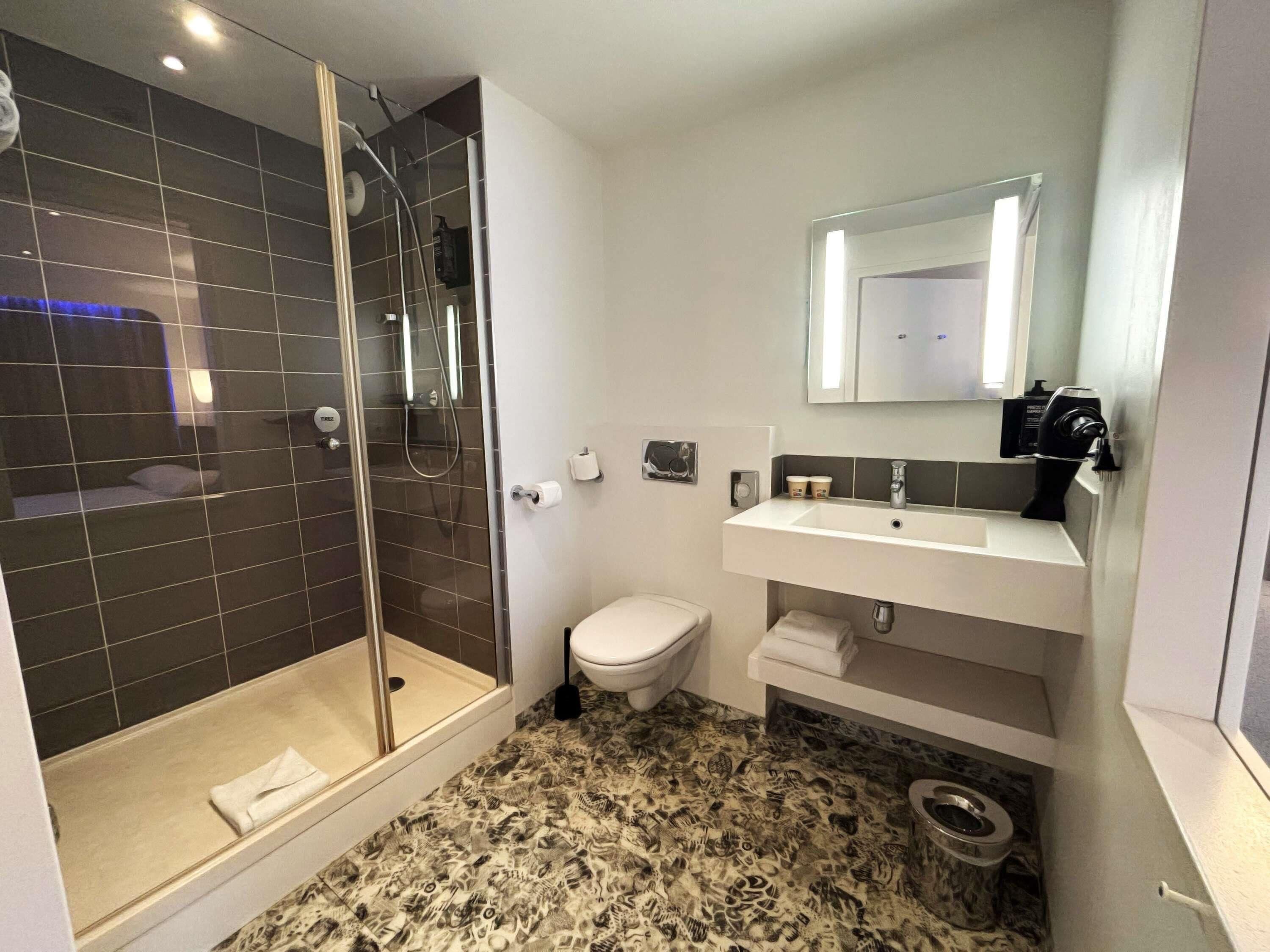 A modern bathroom with a glass-enclosed shower, a toilet, a sink with a mirror, and a patterned floor.
