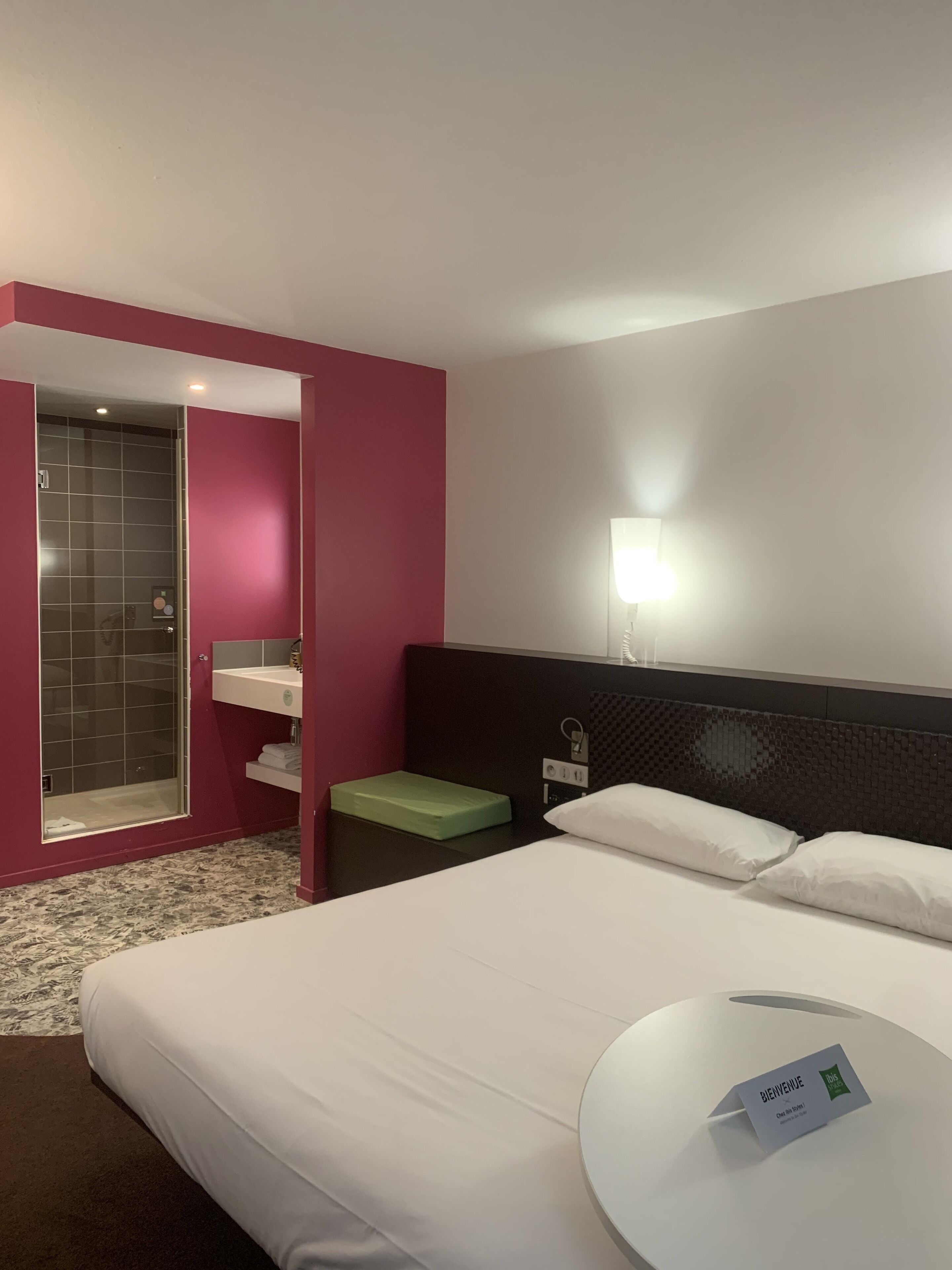 A hotel room with a bed, a bedside table, a nightstand, a mirror, a wall-mounted light, and a bathroom with a shower and a sink.