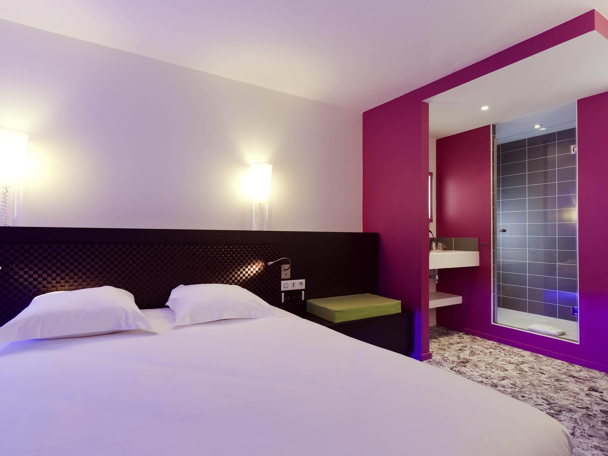 A hotel room with a bed, a nightstand, a wall-mounted lamp, a bathroom with a glass door, and a patterned carpet.