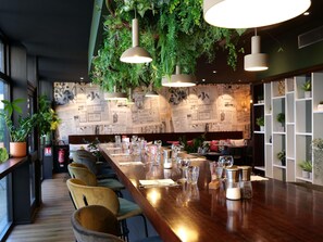 A restaurant with a long wooden counter, multiple chairs, hanging plants, and a wall with newspaper clippings. - ibis Styles Lorient Caudan (Caudan)
