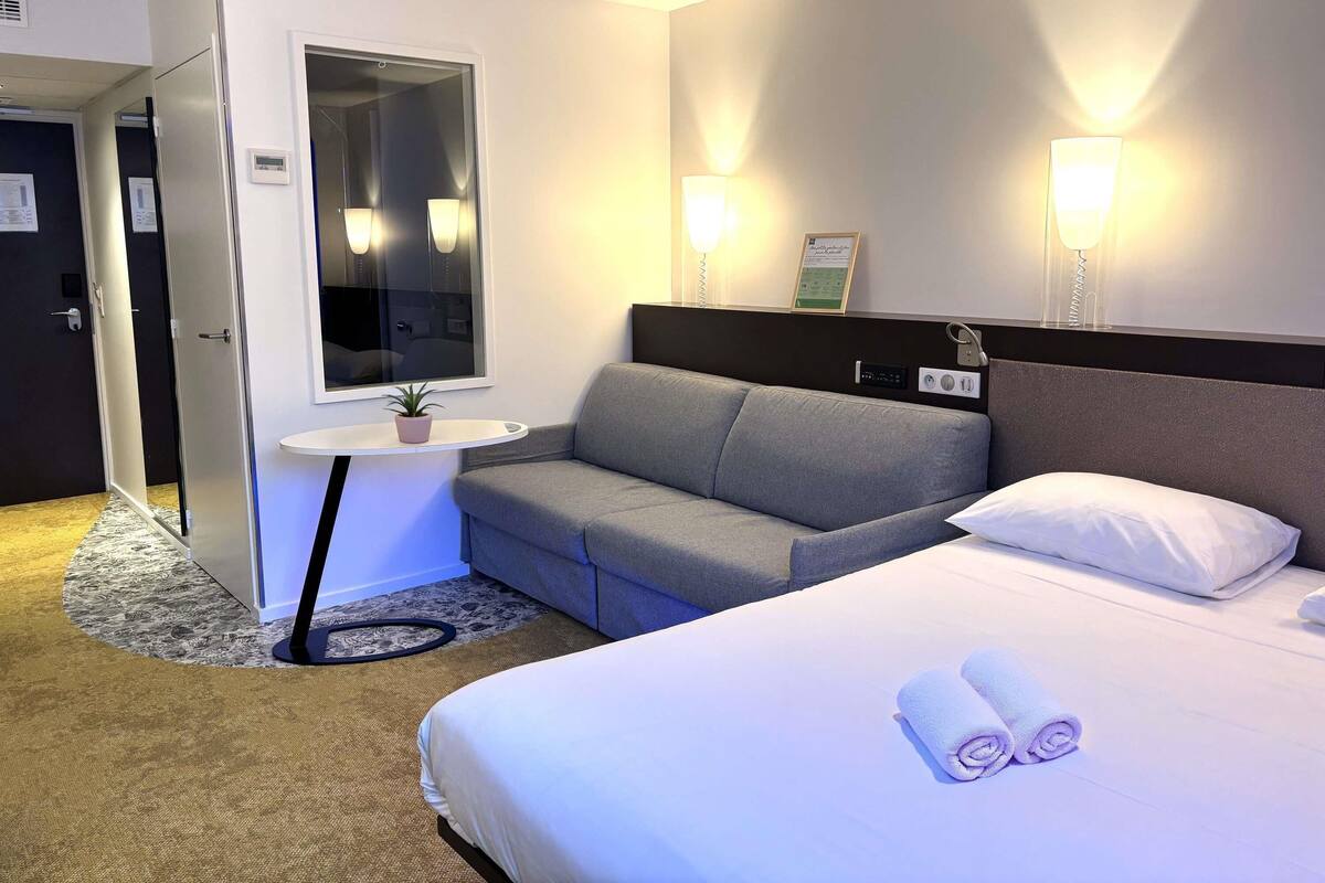 A hotel room with a bed, a sofa, a small table with a plant, a mirror, and wall-mounted lights.
