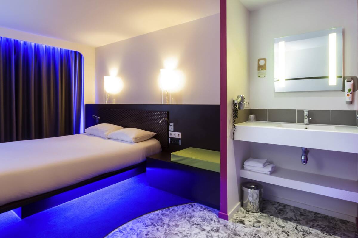 A hotel room with a bed, bedside lamps, a nightstand, a bathroom with a sink and mirror, and a trash can.